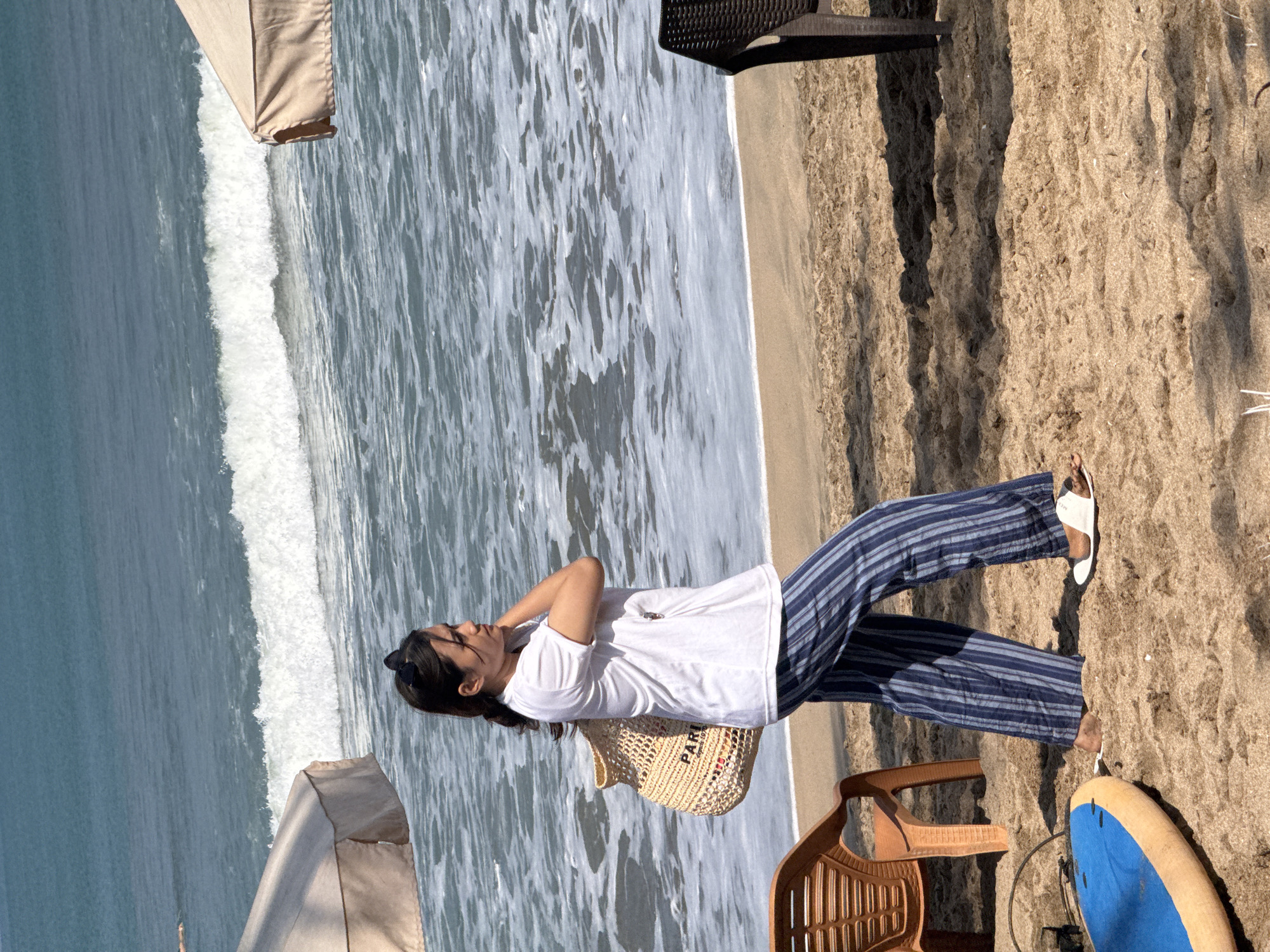 A woman walks along the sandy shore of Kuta beach in Bali, Indonesia, on a sunny day. She is wearing a white t-shirt, blue and white striped pants, and white sandals. She carries a woven straw bag over her shoulder. In the background, the turquoise ocean waves crash onto the beach, and two beach umbrellas are visible. An orange plastic chair sits on the sand to her left, and a dark-colored chair is to her right. A blue and yellow surfboard lies on the sand in the foreground. The sand is textured with footprints and shadows.