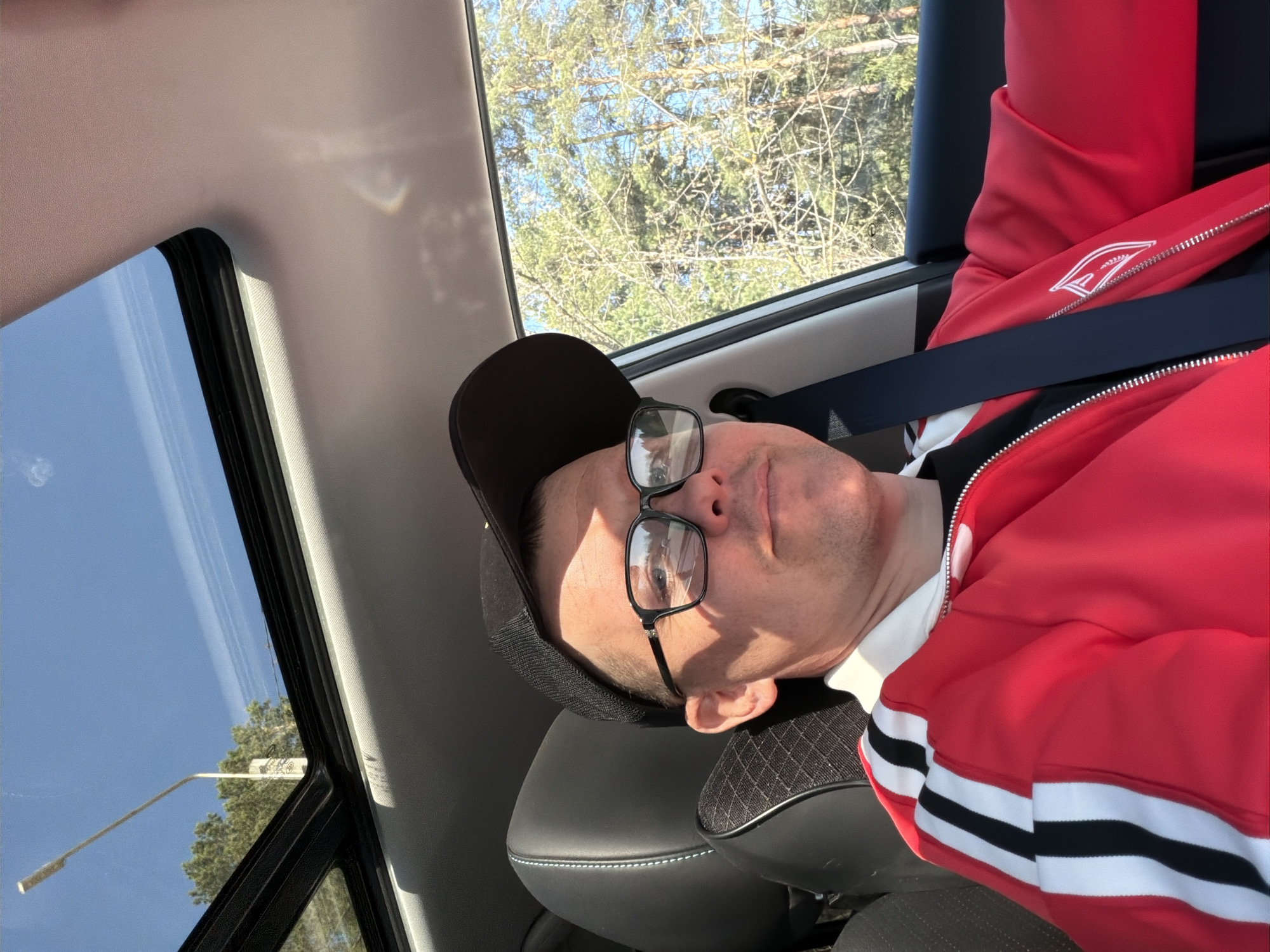 A man in a car is wearing a red jacket with white stripes and a black baseball cap. He has glasses and is wearing a seatbelt. The car has a sunroof. Outside the car, trees and a blue sky are visible. The location appears to be the city of Chaikovsky, Russia.