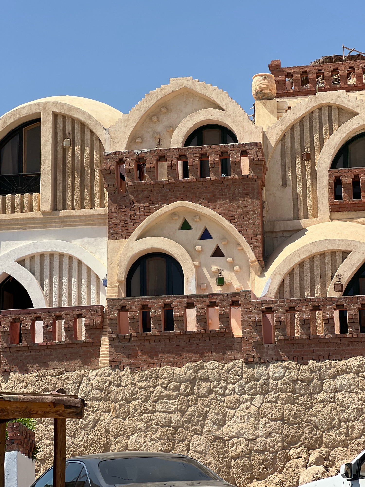 The image shows the exterior of a building with a distinctive architectural style in Sharm El Sheikh, Egypt. The building features curved, domed roofs and arched windows, constructed with a combination of rough stone, brick, and stucco. The lower portion of the building is a textured stone wall. The upper levels display ornate brickwork, including decorative railings and openings. There are small, colorful triangular glass panes embedded in the stucco above one of the arched windows. A large ceramic amphora sits on a ledge near the top of the building. Sunlight illuminates the scene, casting shadows and highlighting the textures of the materials. The sky is clear and blue. A portion of a car is visible in the lower foreground. The image captures the unique architectural aesthetic of the region.