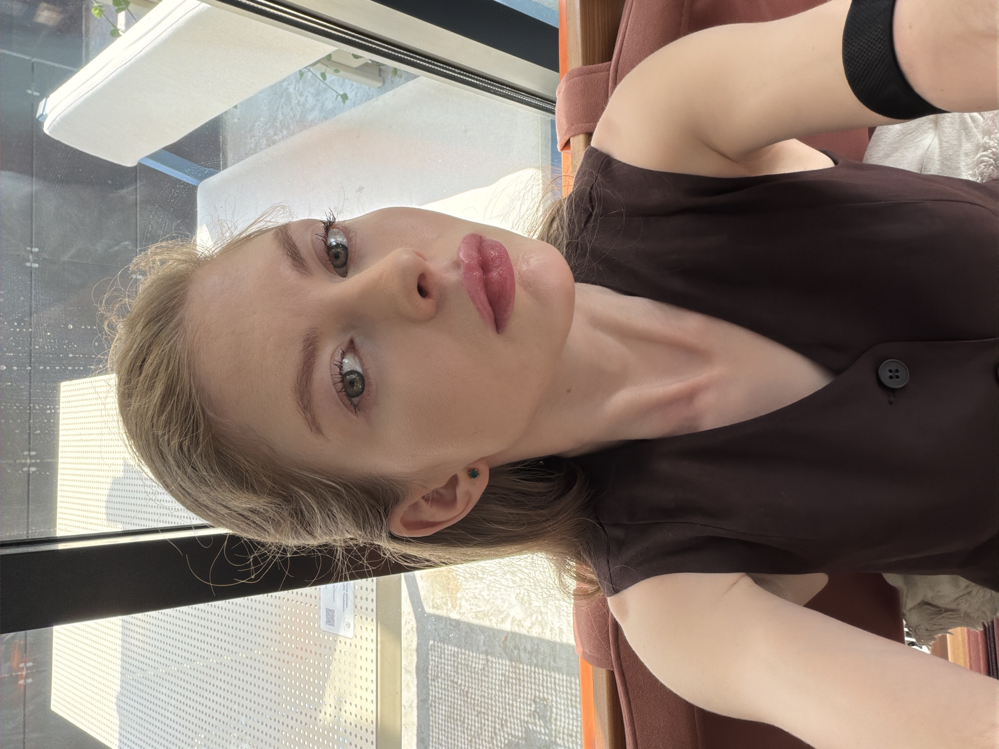 A fair-skinned woman with light brown hair and green eyes is taking a selfie. She is wearing a brown sleeveless top. Her arm is visible with a black wristband. She appears to be indoors, near a large window with a view of an outdoor seating area. The location is potentially in Moscow, Russia. The presence of a window and outdoor seating area are the only possible hints of this being in Moscow Russia.