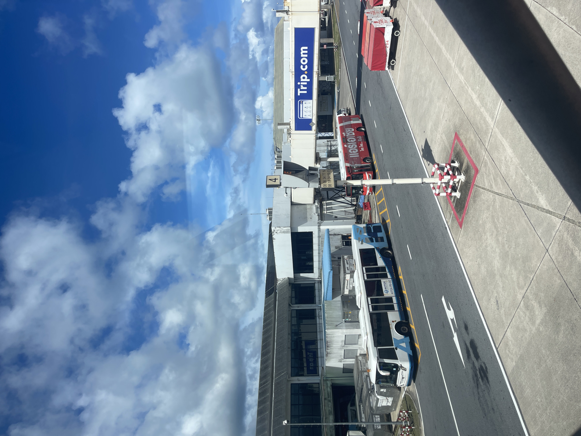 This is a daytime, outdoor, eye-level, full shot of an airport tarmac in Mai Khao, Thailand. The image is dominated by a bright blue sky with scattered white clouds. In the foreground, a concrete walkway is visible, with a diagonal shadow cast across it. The middle ground features an asphalt tarmac with white lane markings and directional arrows. Two buses are present on the tarmac: a white and blue bus on the left, and a red bus with Thai text on the right. Behind the buses, a jet bridge extends from a terminal building. A large blue banner with "Trip.com" logo is displayed on the jet bridge. Several luggage carts, one with a red cover and the number "294" visible, are parked on the right side of the tarmac. The background shows a long, low airport terminal building with dark windows. The overall scene depicts a typical airport operational area under a clear sky.