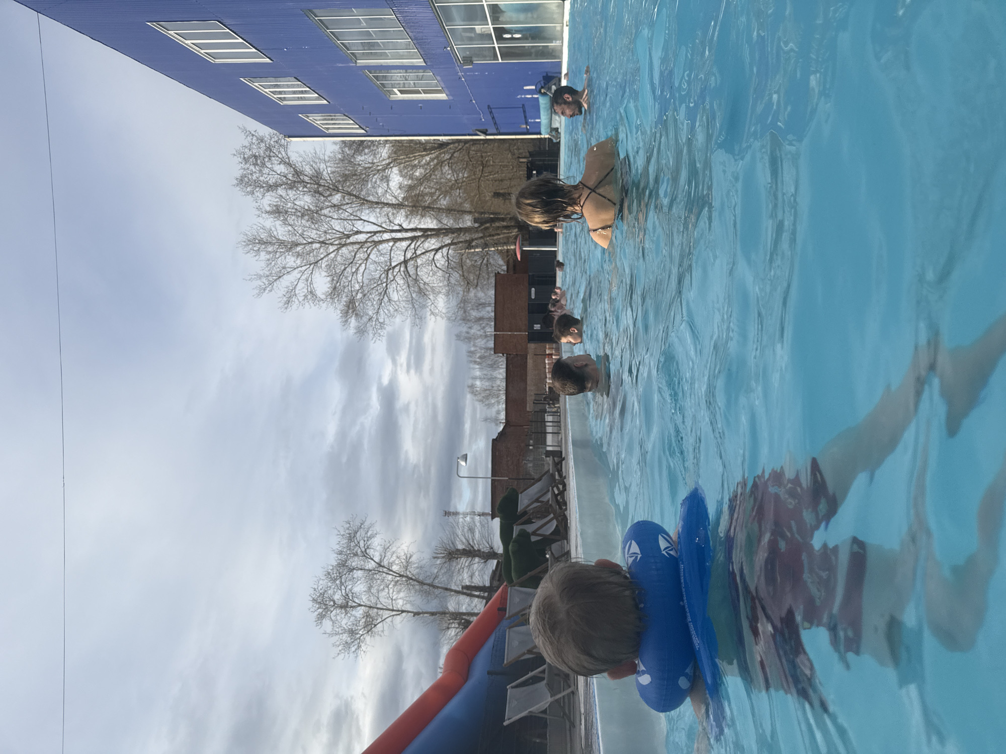 The image, rotated 90 degrees counter-clockwise, depicts an outdoor swimming pool scene under an overcast sky. Several people are visible in the blue water of the pool. In the foreground, a young child with light hair wears bright blue inflatable armbands, and another person's body is partially submerged underwater to the right, wearing patterned swim trunks.
Further back in the pool, a woman with dark hair, seen from behind, wears a dark swimsuit with crisscross straps. To her right and slightly behind, a man with a beard and goggles is visible, with other indistinct figures swimming or standing in the water.
On the left side of the pool, an inflatable structure, partially orange and blue, resembles a slide. Beside the pool, there are several deck chairs or loungers. The background features numerous trees with bare branches, suggesting late autumn, winter, or early spring. A large, prominent blue building with multiple rectangular windows is visible behind the trees and pool area. The sky is largely covered with clouds, indicating daytime. A thin wire is visible stretching across the top left portion of the sky. No discernible text is visible in the image. The scene is set in Glazov, Russia, suggesting this is a heated outdoor pool given the visible season.