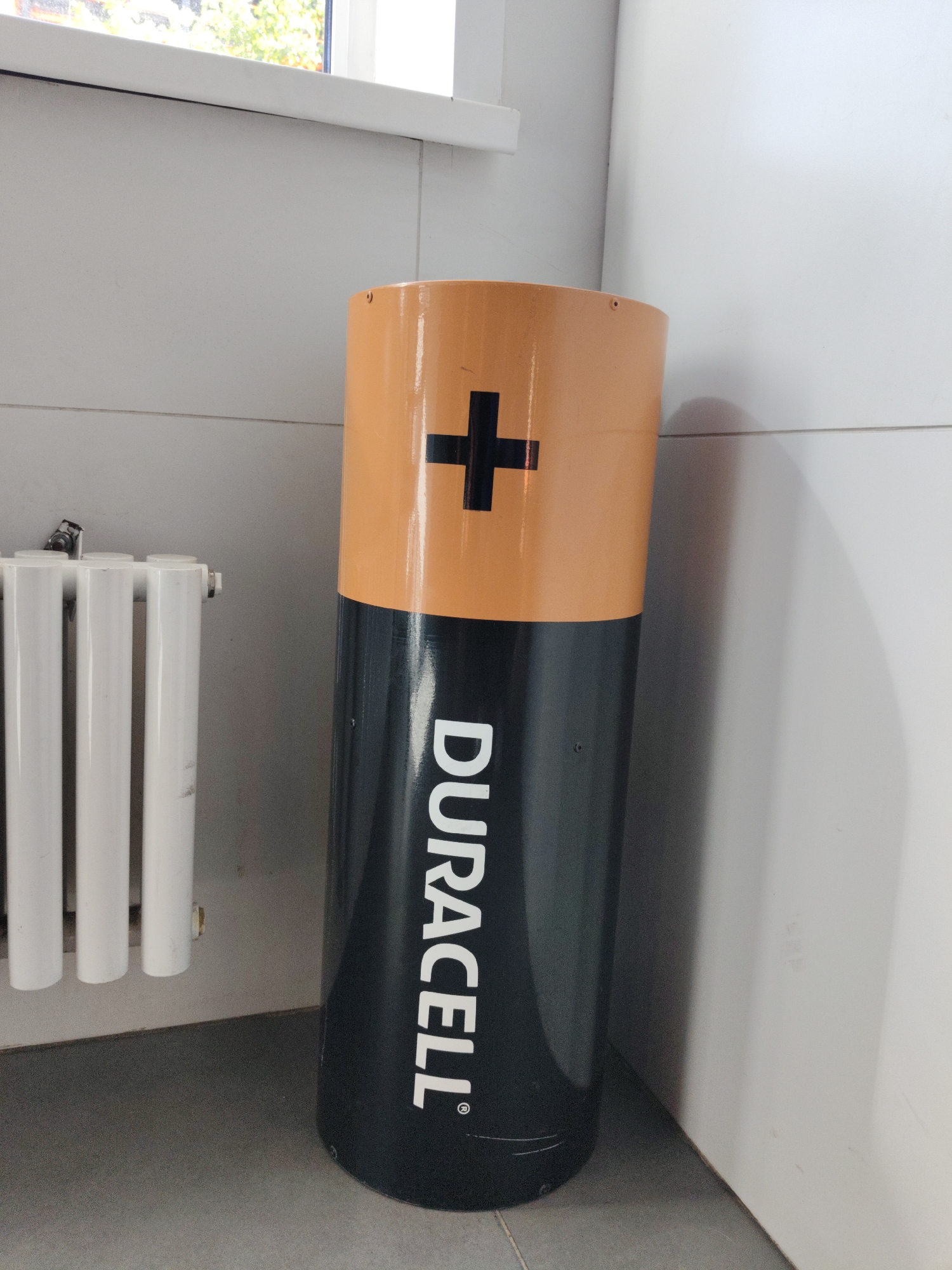 The image shows a large cylindrical object resembling a Duracell battery standing next to a white radiator in what appears to be an indoor setting. The battery-shaped object is predominantly black with an orange top section, and the word "DURACELL" is written vertically in white text on the black part. A black plus sign is visible on the orange top. The radiator is white and has a ribbed surface. The background consists of a light-colored wall, a portion of a window with greenery visible outside, and a grey floor. There are no people or animals in the image. The location is identified as Kommunarka, Russia.