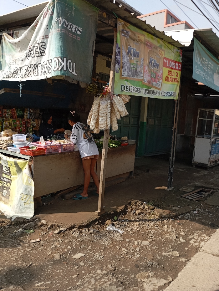 An outdoor vendor stall operates in Cipatik, Indonesia, under natural daylight. The unpaved ground in the foreground is packed dirt and gravel with debris, including plastic litter. A concrete curb is on the far right. A building with a green double door and a gray-roofed structure forms the background, with faint overhead power lines visible top right.
The makeshift stall has a corrugated metal roof on wooden/metal poles, adorned with fabric banners. A large green and white banner over the main counter reads "TOKO SAYURAN" (Vegetable Shop) and "SEDIA." To its right, a yellow and green 'Klin' detergent banner advertises "Rp 1.000 X 2, BELI 3 GRATIS" (Buy 2 for Rp 1,000, Get 3 Free). Another small banner is partially visible far right. Strings of dried/packaged food, possibly snacks, hang from a central pole.
Behind the counter, a person in a dark top (possibly headscarf) engages with items, facing right. In front, a second person, likely female, in a white t-shirt, white shorts, and light blue flip-flops, leans over the counter towards the first, indicating interaction.
The robust counter, likely wood or concrete, holds plastic containers, a red basket, and produce/packaged goods. Shelves of packaged snacks are to the far left inside the stall. A pile of green vegetables is to the counter's right, and a white display unit/refrigerator stands against the building wall.