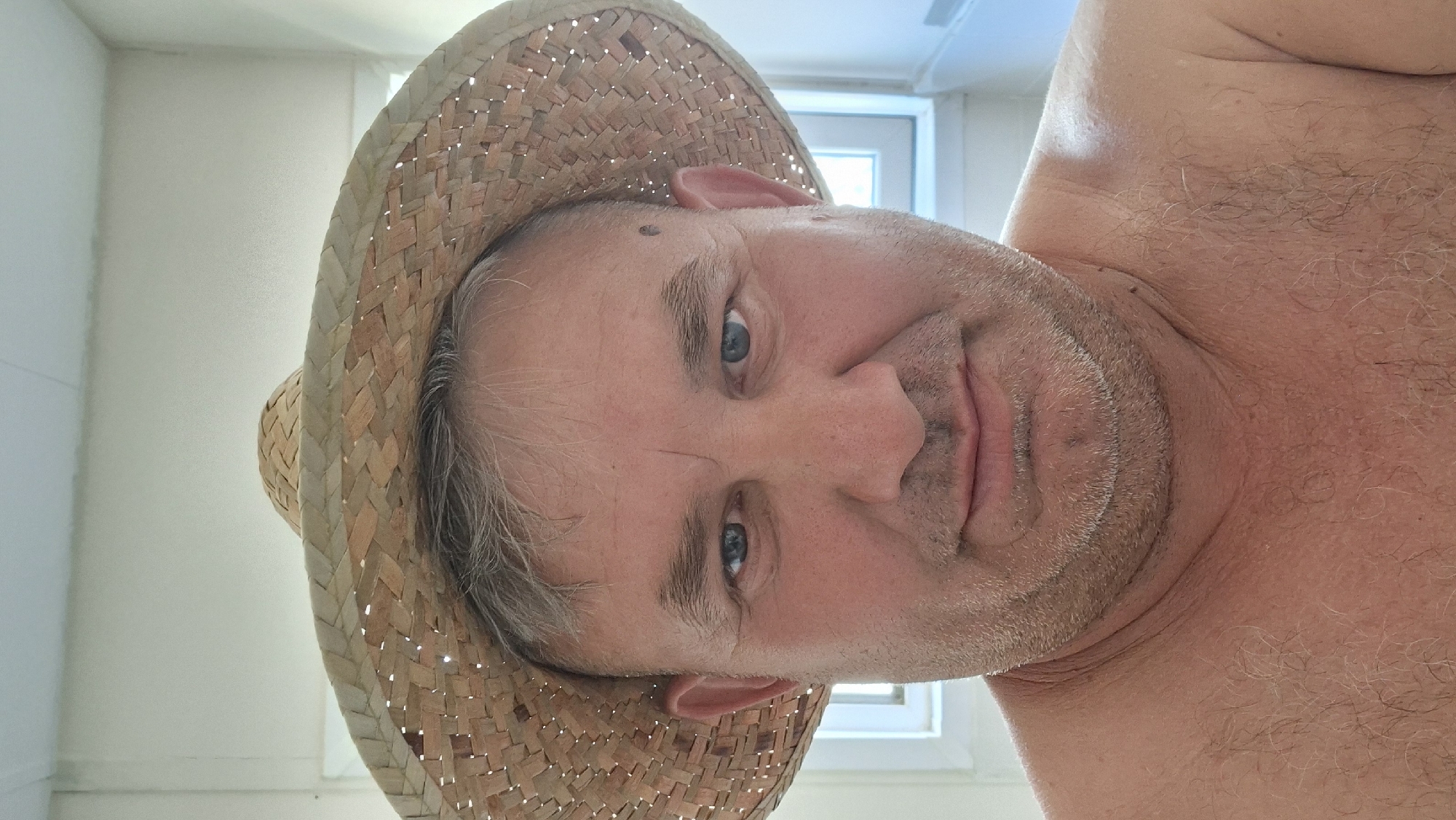 A close-up, head-and-shoulders selfie of a middle-aged man wearing a straw hat. He has graying hair, blue eyes, and a light beard and mustache. His bare chest is visible. The background appears to be indoors, with a window and white walls. The image was taken in Hîncești, Moldova.