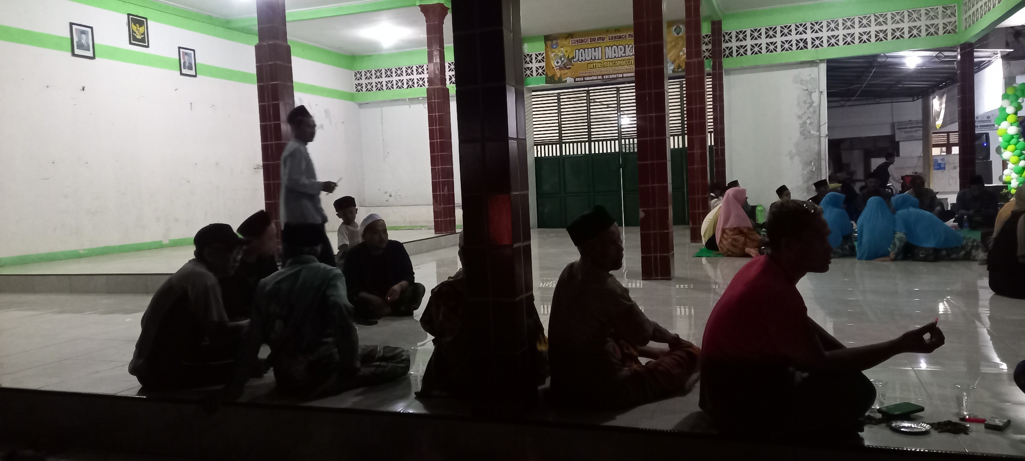 The image depicts an indoor gathering of people, likely at a community hall or mosque, given the architectural style and the presence of religious imagery. The location is identified as Sidamulya, Indonesia. The scene is dimly lit, with fluorescent lighting from the ceiling.
In the foreground, several individuals are seated on the floor, some facing away from the camera and others in profile. One man in the lower right foreground wears a red shirt and is holding a small object, possibly a cigarette or incense, between his fingers. He is looking towards the left. Next to him are a glass, a small plate, and what appears to be a mobile phone. To his left, another person in a brown garment is seated with their back to the camera, their head adorned with a black cap.
In the mid-ground, a group of people, including men and women wearing headscarves in various colors (pink and blue), are also seated on the floor. A child in a white shirt and black cap is visible in the center of the mid-ground, standing near an adult male in a light-colored shirt and black cap, who appears to be walking and holding a small object.
The background features a white wall with a green stripe running horizontally. Three framed pictures hang on the wall, one of which appears to be a portrait. Large, reddish-brown tiled pillars are strategically placed throughout the space, supporting the ceiling structure. Further in the background, a large green gate or door is visible, along with a banner that has text in Indonesian, including the words "JAUHI NARK" which suggests an anti-drug campaign. The overall atmosphere appears to be a communal gathering or event.