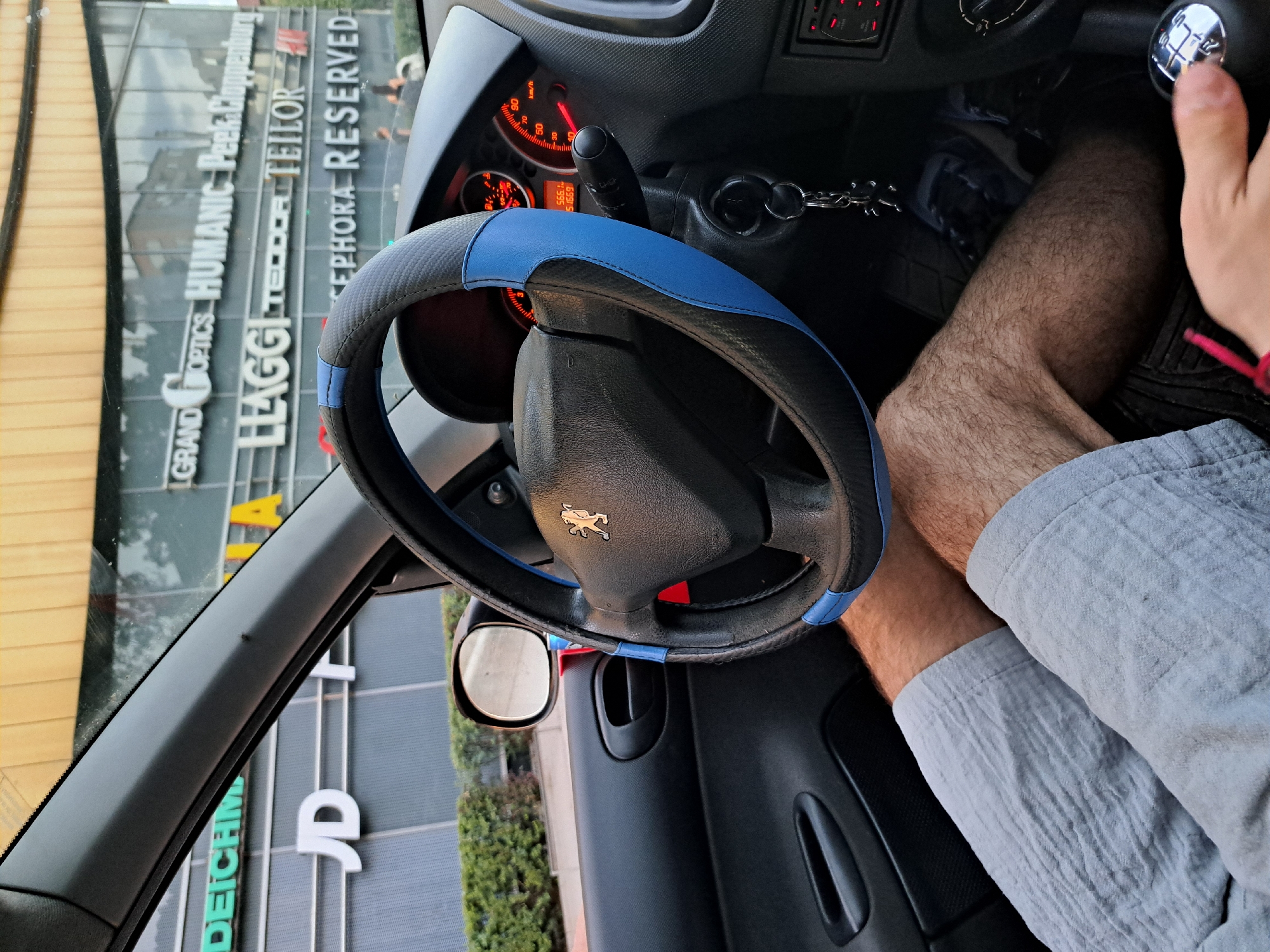 The image shows the interior of a car, specifically the driver's side. A Peugeot steering wheel with a blue and black cover is visible. The dashboard and parts of the center console are also in view. A person's leg and part of their arm are present in the lower right, suggesting the driver is in the car.
Through the car window, parts of buildings with signage are visible. The signs read: "DEICHMANN," "GRAND OPTICS HUMANIC Peek & Cloppenburg," "LAGGI TEODOR TEILOR," and "SEPHORA RESERVED." Based on the signs, the location is most likely in the city of Sofia, Bulgaria, where these brands have a presence.