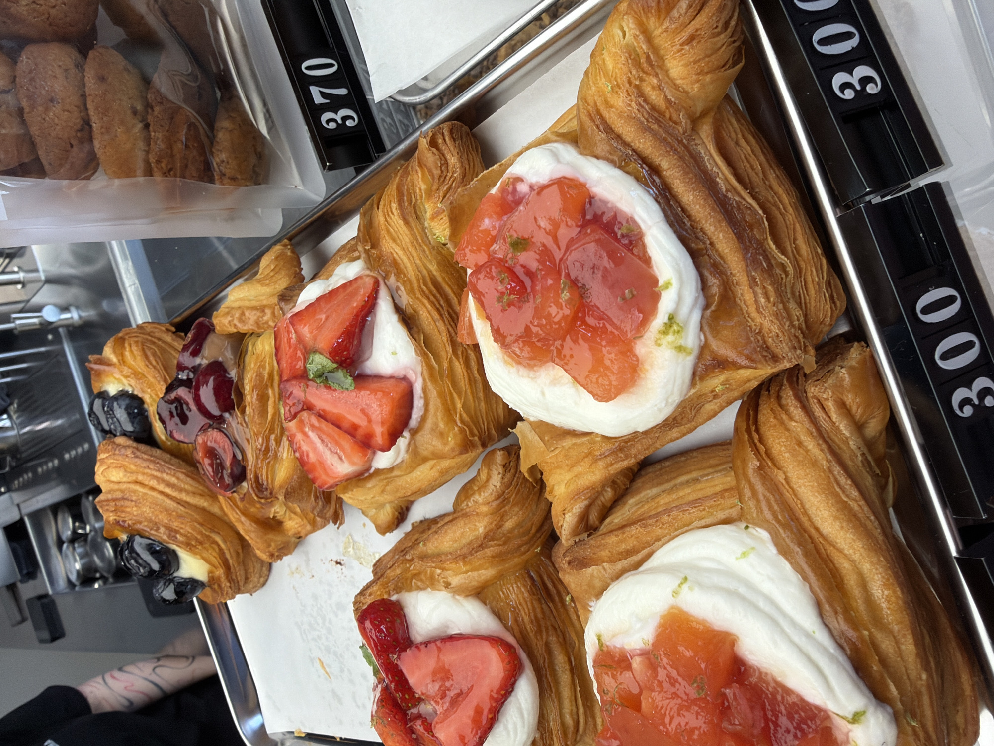 The image depicts a close-up view of several pastries, likely danishes, displayed on a metal tray. The pastries are golden brown and flaky, with various toppings. Some are topped with fresh strawberries and a dollop of white cream, while others feature whole blueberries and cherries, also with cream. A few pastries appear to have a strawberry compote or jam topping, garnished with what looks like lime zest. The pastries are arranged on a white paper liner on the tray. In the background, a blurred image shows a kitchen or service area with stainless steel appliances. Black price tags with white numbers are visible along the bottom edge of the tray, indicating prices of "370" and "300". The overall scene suggests a bakery or cafe setting. The image was taken in Izhevsk, Russia.