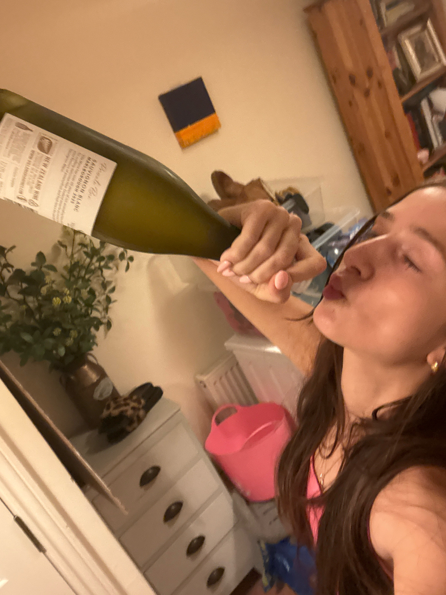 The image depicts a woman tilting a bottle of white wine towards her lips, with her eyes closed. She is wearing a pink top and a necklace, with her long, dark hair falling around her shoulders. A patterned scarf or cloth is visible on a chest of drawers in the background, along with a vase of artificial greenery. The scene appears to be indoors, possibly a domestic setting. There is no information in the image that places it in the city of Reigate and Banstead, United Kingdom.
