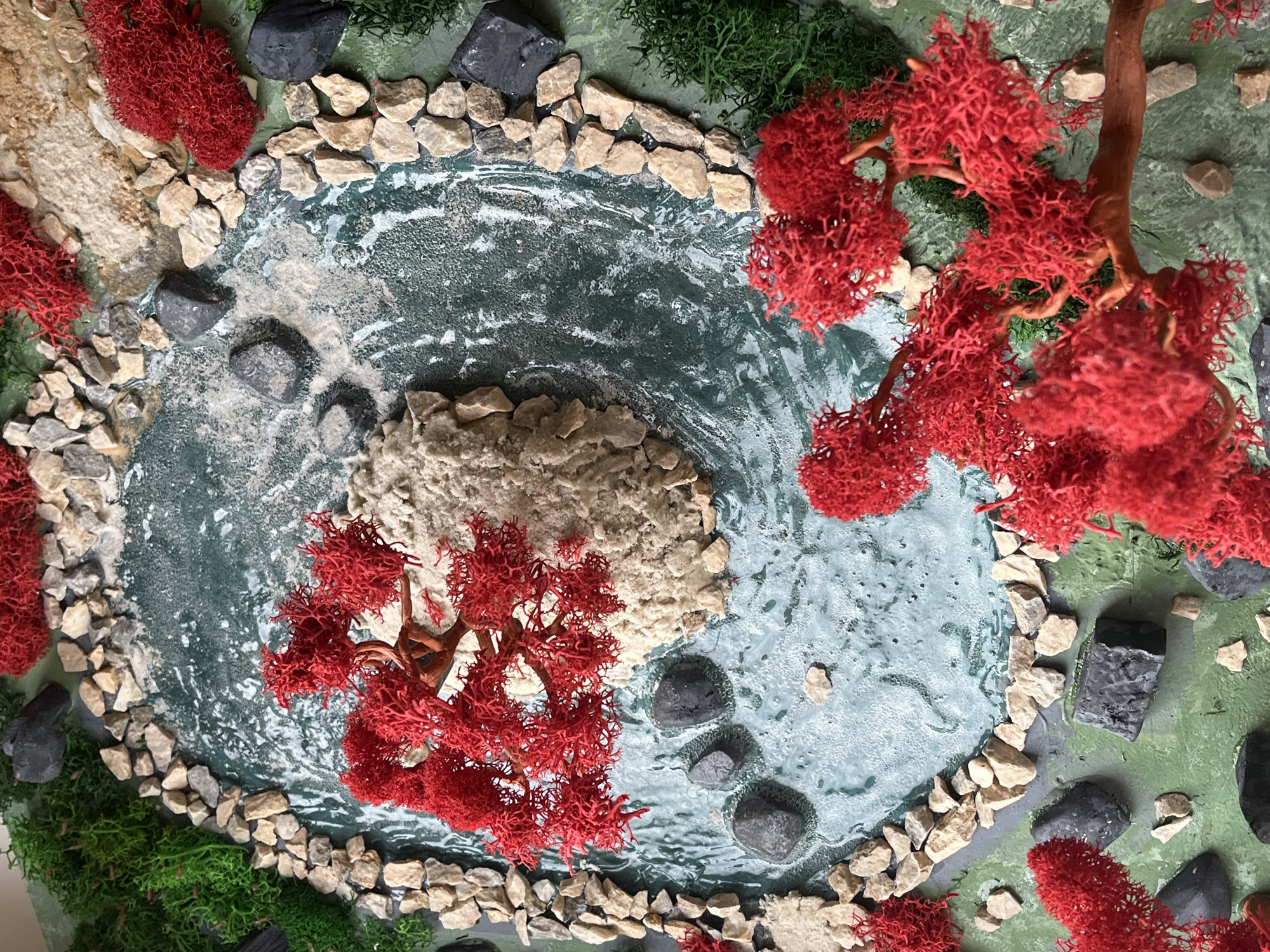 The image depicts a miniature diorama of a pond bordered by rocks and foliage. The water is simulated with a textured blue-green surface, an island of beige material in the center. Red faux trees surround the pond and green mossy areas frame the landscape. Black rocks are scattered around the scene. There are no people depicted in the diorama. The location in the city of Saint Petersburg, Russia is not discernible from the image.