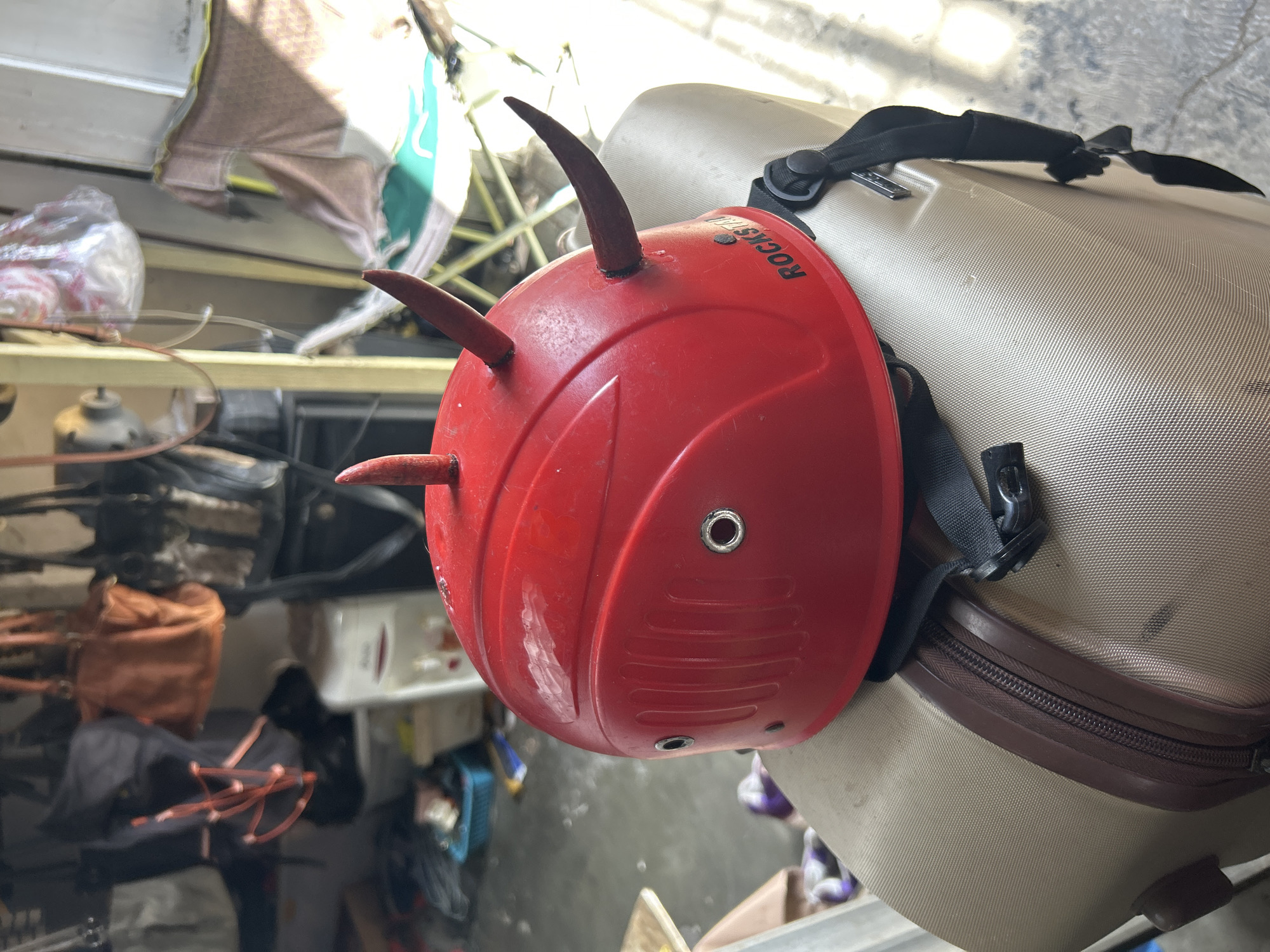 A close-up shot focuses on a red novelty helmet with two small red horns, resembling a devil's horns, attached to its top. The helmet has two visible metal eyelets on its sides and a ribbed texture on the front. The word "ROCKSTAR" is printed in black lettering on the right side of the helmet. The helmet is resting on top of a tan suitcase with a brown zipper and trim. A black strap with a buckle is attached to the helmet and extends down the side of the suitcase. The background is out of focus but appears to be an indoor or covered area with various items like bags, boxes, and a white appliance. The lighting suggests it is daytime, with sunlight casting shadows and illuminating parts of the scene. The location is identified as Denpasar, Indonesia.