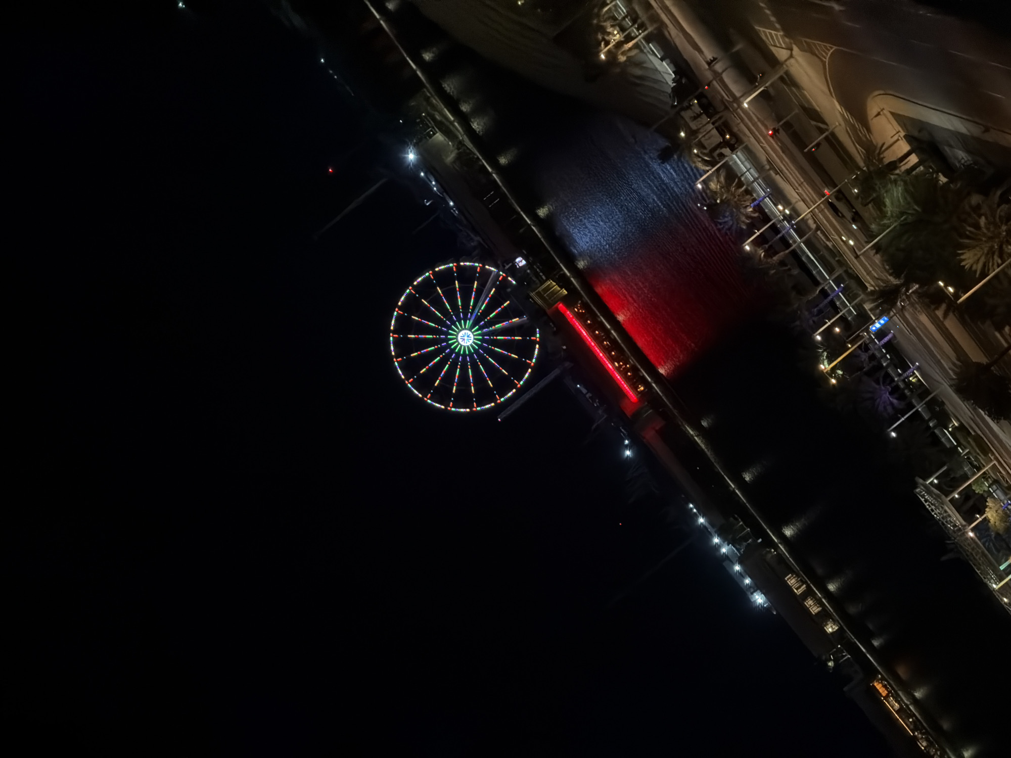A high-angle nighttime view of a waterfront area in an unknown city shows a Ferris wheel illuminated with colorful lights. The Ferris wheel is centrally located in the image, with its bright lights contrasting against the dark sky. Below the Ferris wheel, a body of water reflects the lights from the surrounding buildings and the Ferris wheel itself. Along the edge of the water, a promenade or walkway is visible, lined with lights and palm trees. Further in the foreground, a street with traffic lights and cars is present, also illuminated by streetlights. The scene suggests a lively urban environment, likely a tourist or entertainment district.