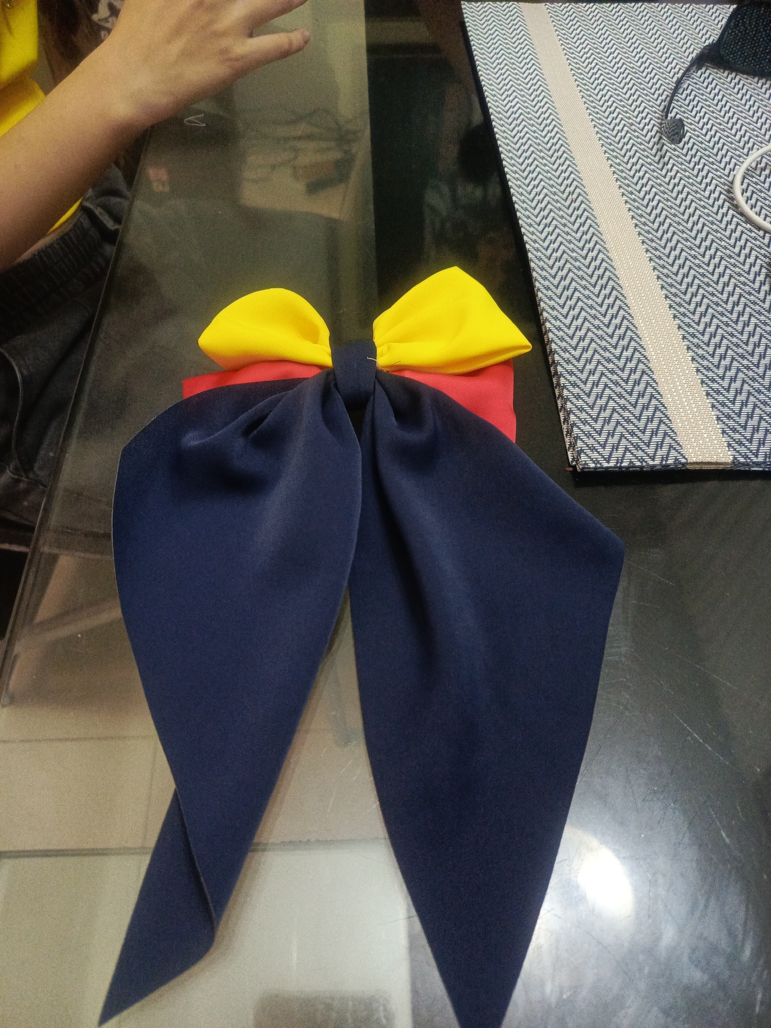 A close-up, overhead view shows a handcrafted bow made of dark blue, red, and yellow fabric. The bow is the central subject, positioned on a dark, reflective surface that appears to be a table. To the right, a patterned textile, possibly a placemat or runner, is visible. In the upper left corner, a person's arm and hand are partially in frame, suggesting they are either making or arranging the bow. The lighting is direct, casting some reflections on the table surface. The image quality is moderate, with some graininess. The location is not determinable from the visual elements within the image.