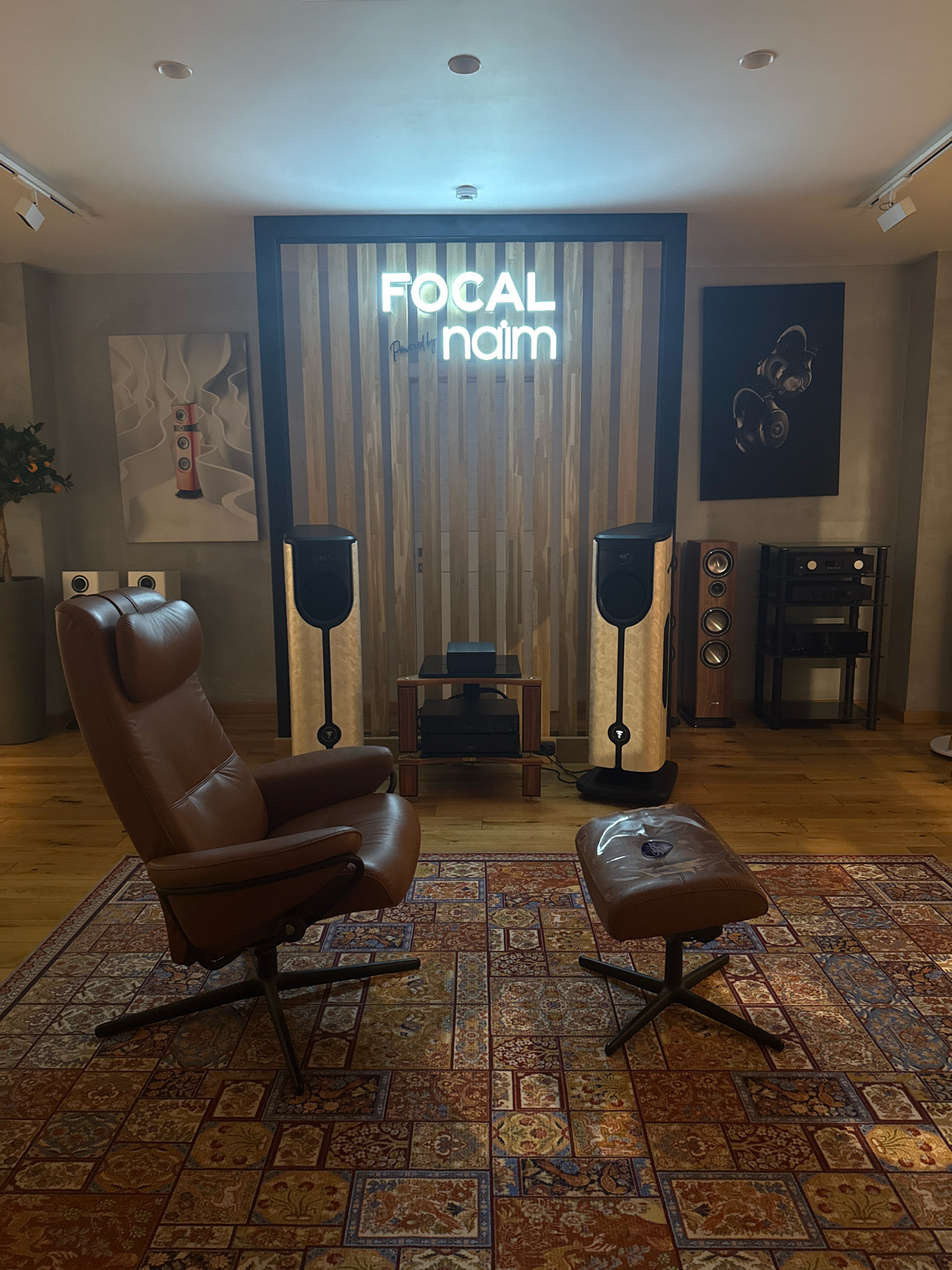 The image shows a well-lit room with high-end audio equipment. In the foreground, a brown leather armchair and ottoman are placed on an ornate, colorful rug. Behind the armchair, two large, modern speakers with light-colored wood veneer and black accents stand tall. To the right of these speakers, a smaller, darker wood speaker is visible, and further back, a multi-tiered black rack holds electronic components. A neon sign with the words "FOCAL naim" in white is mounted on a wooden slatted wall in the background. To the left of the neon sign, a framed abstract artwork depicting a red cylindrical object is displayed. On the far right, another framed artwork shows a pair of black headphones against a dark background. The room has polished wooden floors and a neutral-colored wall. The overall ambiance suggests a showroom or a dedicated listening room. The location is not specified in the image and cannot be determined.