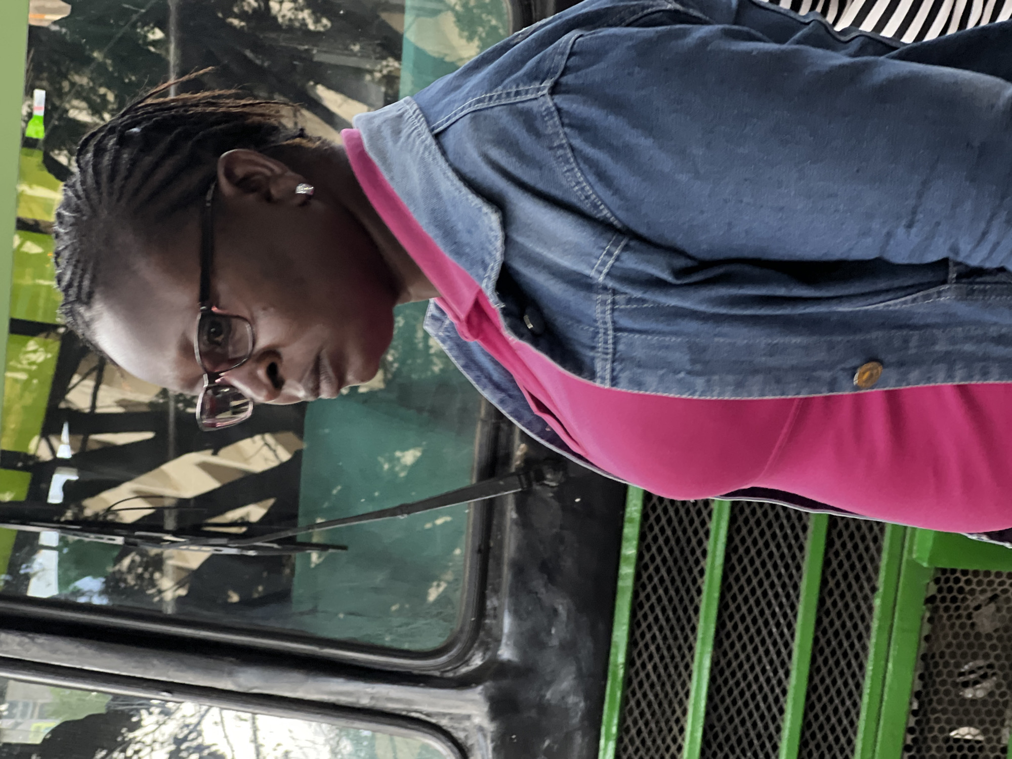 A woman with dark skin and braided hair wearing glasses, a pink top, and a denim jacket is standing in front of a green bus. The bus appears to be in Nairobi, Kenya. The woman is looking down, and part of a black and white striped garment is visible in the background. The bus has windshield wipers and a green grill.
