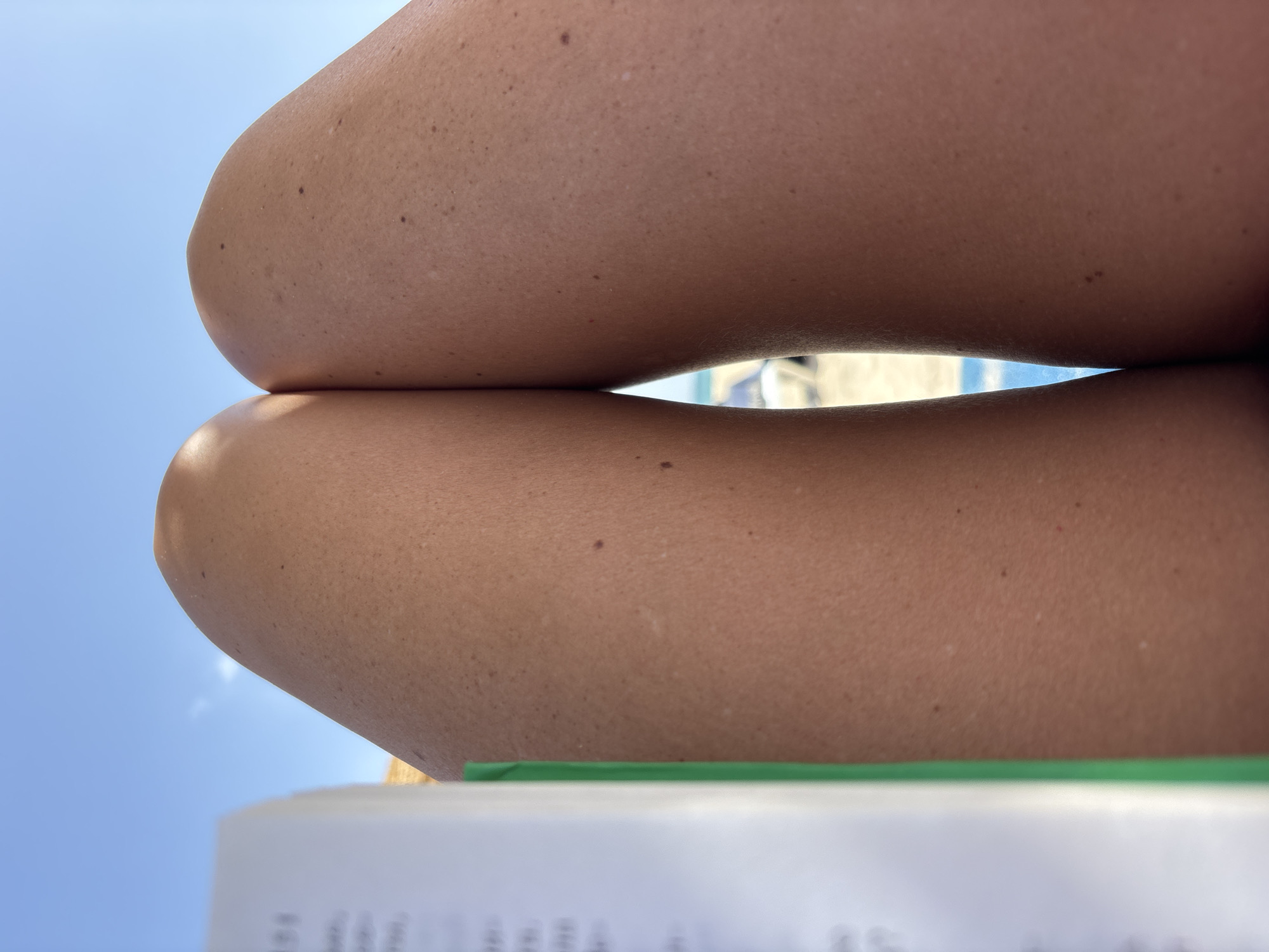 A close-up, low-angle, point-of-view shot shows a person's tanned legs resting against a bright blue sky. A partially opened book with a green cover is in the foreground on the left, obscuring a portion of the left leg. The skin on the legs shows a light scattering of freckles and a few small moles. In the background, out of focus, the turquoise water of a swimming pool or the ocean is visible, along with what appears to be beach umbrellas. The scene suggests a relaxed setting, likely outdoors in a warm climate, potentially in Tavira, Portugal, known for its beaches and coastal resorts.