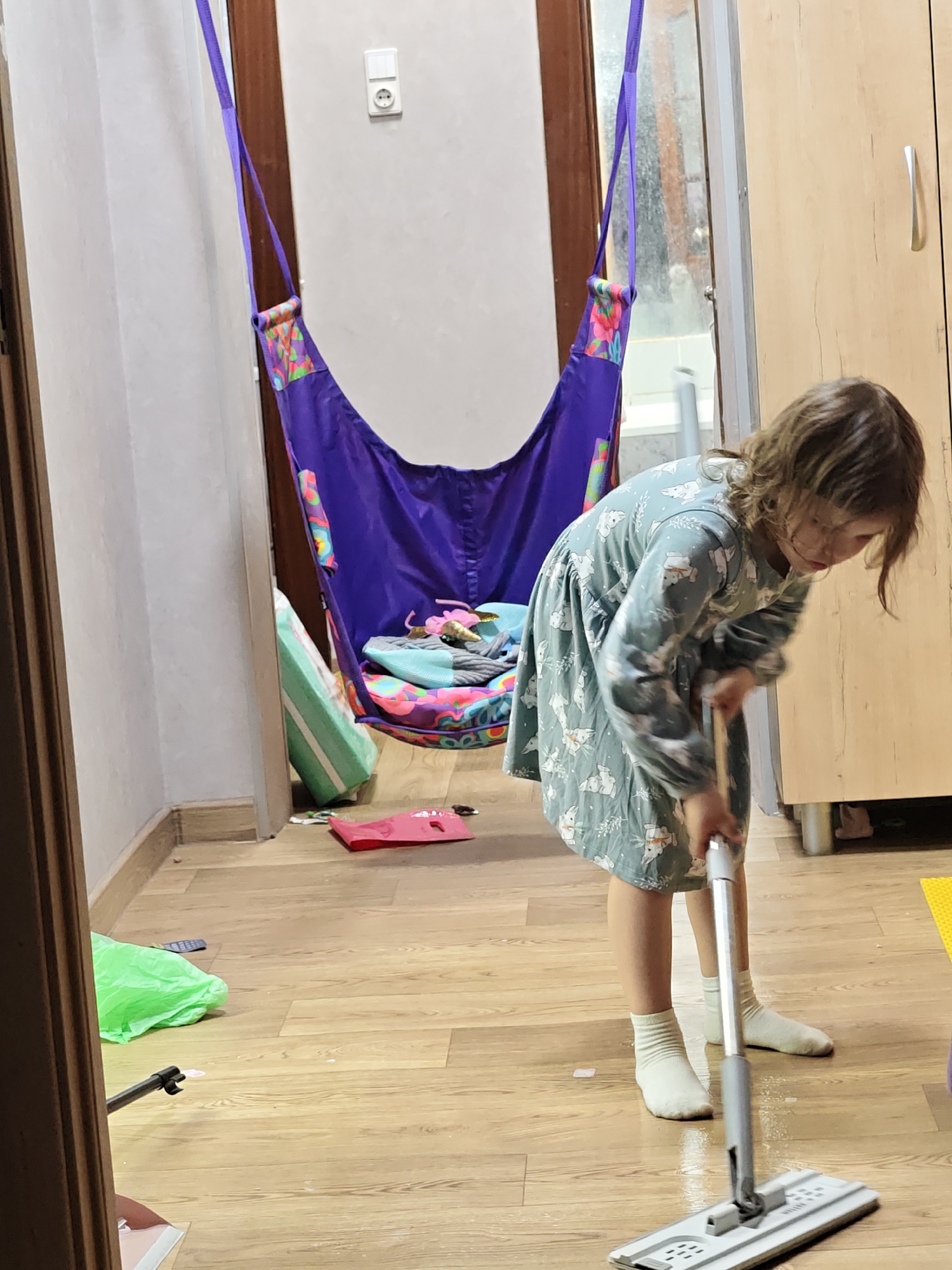 An indoor, eye-level photograph captures a young girl diligently mopping a light wooden laminate floor. The girl, positioned on the right side of the frame, is wearing a long-sleeved, light blue dress patterned with white animals. She has light brown hair and is wearing white socks. She is bent forward, focused on pushing a grey flat mop across a wet section of the floor.
In the background, to the left, a purple fabric swing or hammock hangs from a doorway or frame. The swing features colorful, patterned accents and holds several items, including a patterned cushion and other small belongings, suggesting a child's space. A light-colored wall with a dark wooden door frame is visible on the far left. On the wall behind the swing, a white electrical outlet is mounted. A window showing blurred outdoor elements can be seen between the swing and a light brown wooden wardrobe or cabinet located on the far right.
On the floor in the foreground, near the bottom left, lies a crumpled green plastic bag, and a red paper-like item is closer to the base of the swing. The setting appears to be a typical domestic environment, possibly a child's room or a living area, in Chaikovsky, Russia. The natural light suggests it is daytime. The image conveys a sense of everyday activity with a child engaged in household chores. No discernible text is visible within the frame.