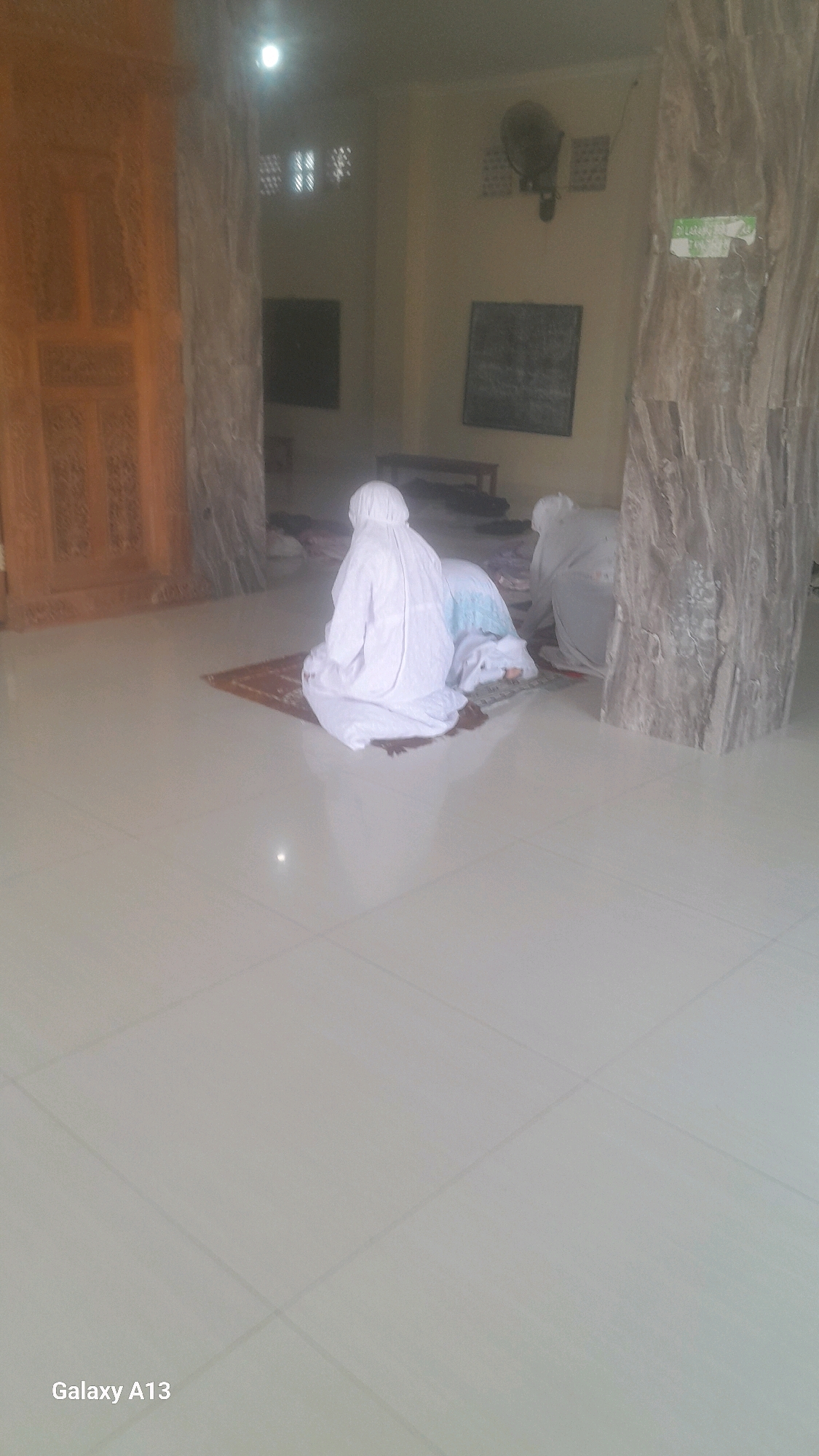 The image depicts the interior of a prayer room, likely a mosque, in Ngelo, Indonesia. Two individuals are observed in the act of prayer, both wearing full white protective suits. One individual is kneeling on a prayer mat, with their head bowed. The second individual is positioned behind and to the right of the first, also in a kneeling posture, with their head lowered. The environment appears to be a simple room with tiled flooring. In the background, a wooden door with intricate carvings is visible to the left, and a wall with a fan and some framed items hangs on the right. The lighting is somewhat dim, with a bright light source visible at the top center of the image. The overall scene suggests a religious gathering taking place within this interior space.