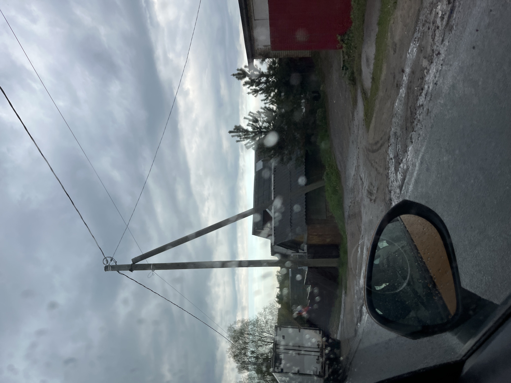 The image, likely taken from inside a vehicle in Ширяево, Russia, captures a rainy day scene. Visible through the rain-streaked windshield and the side mirror is a view of a rural landscape. A utility pole with wires is prominent on the left side. Adjacent to the road, there are structures including a red building with a brick foundation and a dark, barn-like structure with a grey roof. A white box truck is parked further down the road. The road surface is wet and appears to be partially unpaved with mud and puddles along the edge. The sky is overcast.