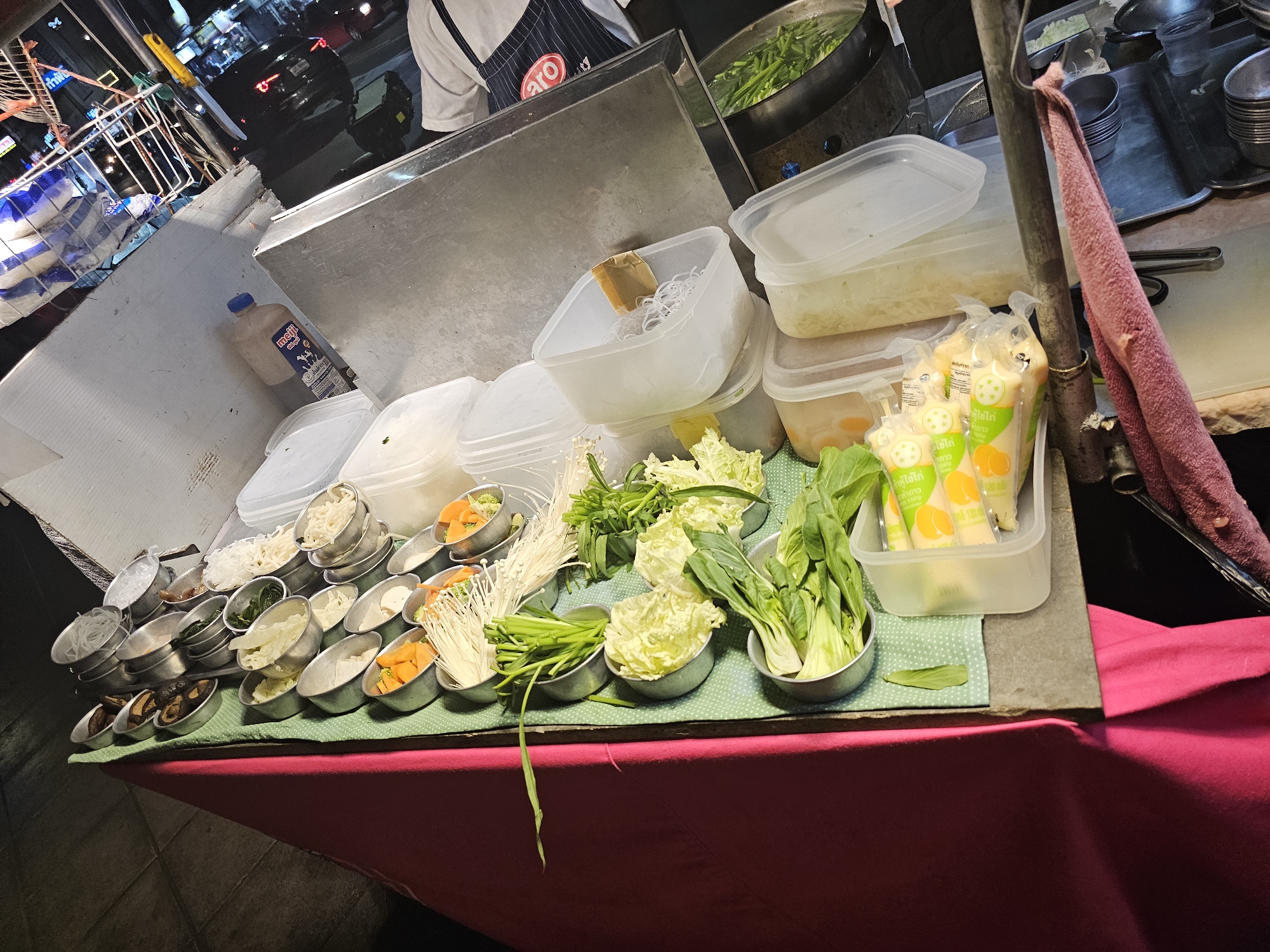 This image depicts an outdoor food stall in Bangkok, Thailand, likely at night given the illumination and background activity. The stall is set up along a street, with a long table laden with various ingredients. The table is covered with a green, polka-dotted cloth and a bright pink cloth draped over its edge.
On the table, an array of metal bowls contains fresh ingredients, including leafy green vegetables like bok choy and napa cabbage, as well as enoki mushrooms and what appear to be diced carrots or pumpkin. There are also clear plastic containers holding noodles and other food items. A bottle of "Meiji" brand milk is visible on a metal counter behind the table. In the background, the busy street scene of Bangkok is evident, with illuminated signs and passing vehicles. A person wearing a white shirt and a dark apron with the "ARO" logo is partially visible to the right, suggesting they are operating the stall. The overall impression is of a vibrant, bustling street food environment.