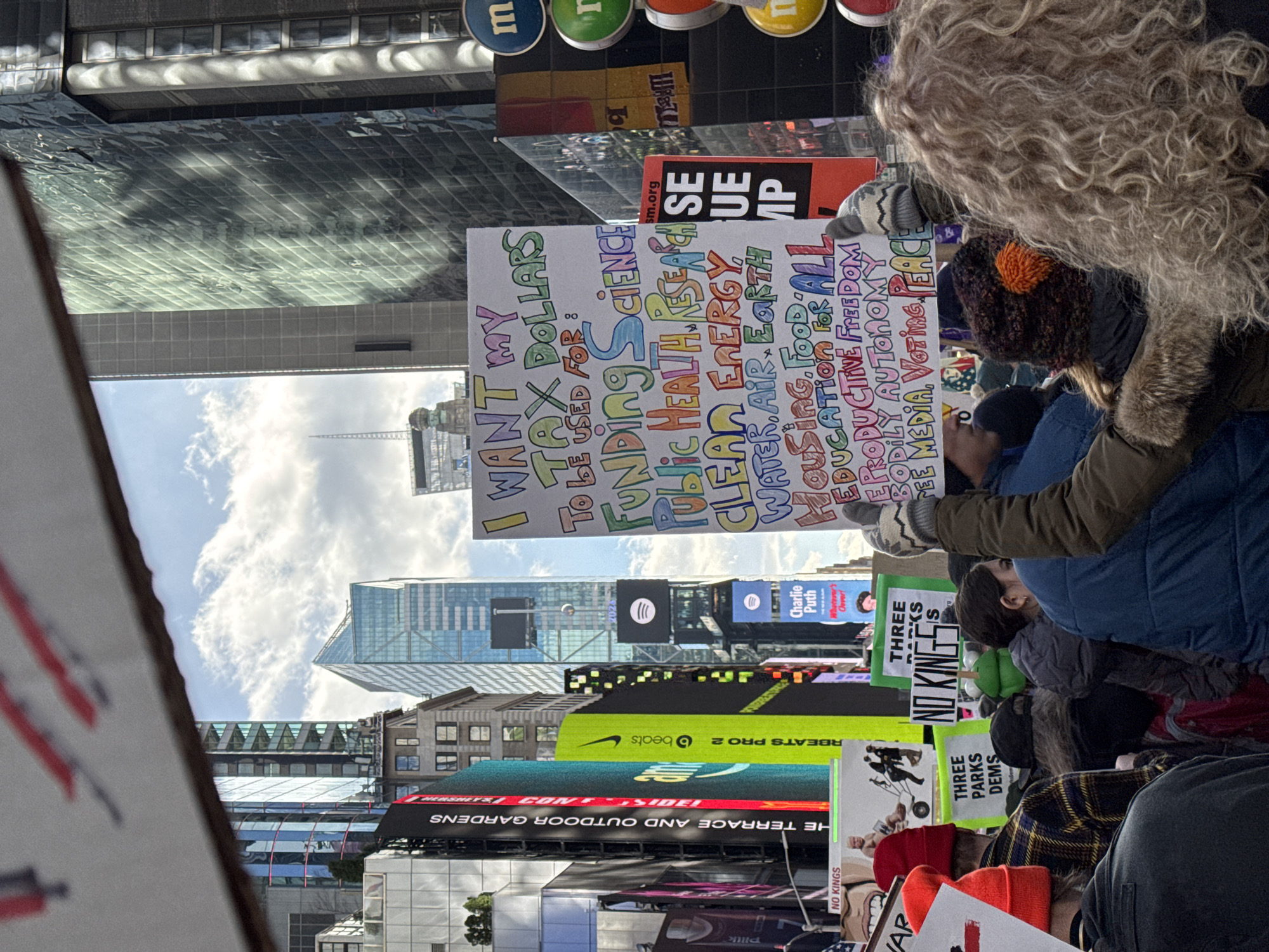 The image depicts a protest in Times Square, New York City. Demonstrators are gathered, holding signs with messages advocating for tax dollar allocation towards science, public health, clean energy, water, air, earth, housing, food, education, reproductive freedom, bodily autonomy, free media, voting, and peace. The scene is surrounded by the vibrant billboards and skyscrapers of Times Square.