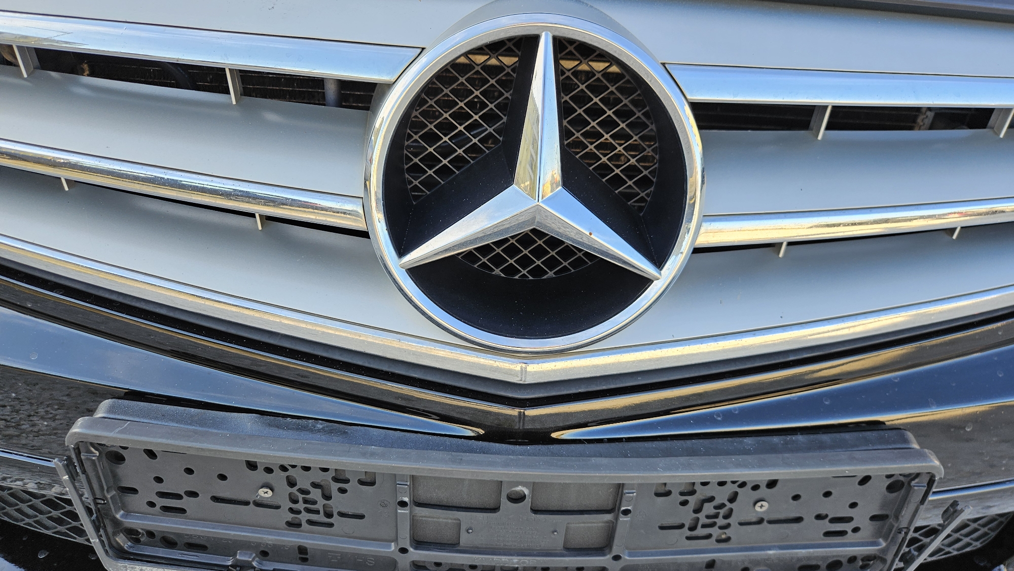 The image shows the front of a Mercedes-Benz car. The prominent feature is the Mercedes-Benz logo, a three-pointed star within a circle, mounted on the car's grill. The grill is silver-colored with horizontal bars. Below the grill, the car's bumper is visible, with a black license plate frame attached. The background is out of focus, preventing identification of a specific location in Stara Zagora, Bulgaria. No people, specific actions, or interactions are discernible in the image. The scene is a close-up view of the car's front end.