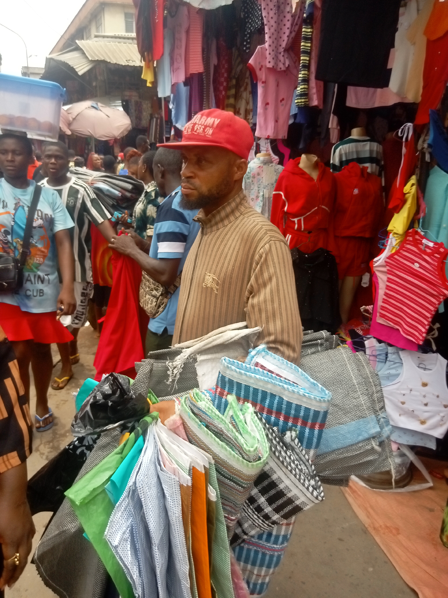 The image depicts a busy market scene in Fegge, Nigeria. In the foreground, a middle-aged man with a beard wears a red cap and a striped brown shirt. He is carrying a large assortment of folded fabrics and clothes in various colors and patterns. Behind him, a crowd of people is visible, some looking at merchandise displayed on racks. The stalls are filled with a variety of clothing, including shirts, dresses, and sportswear. The market environment appears crowded and lively.