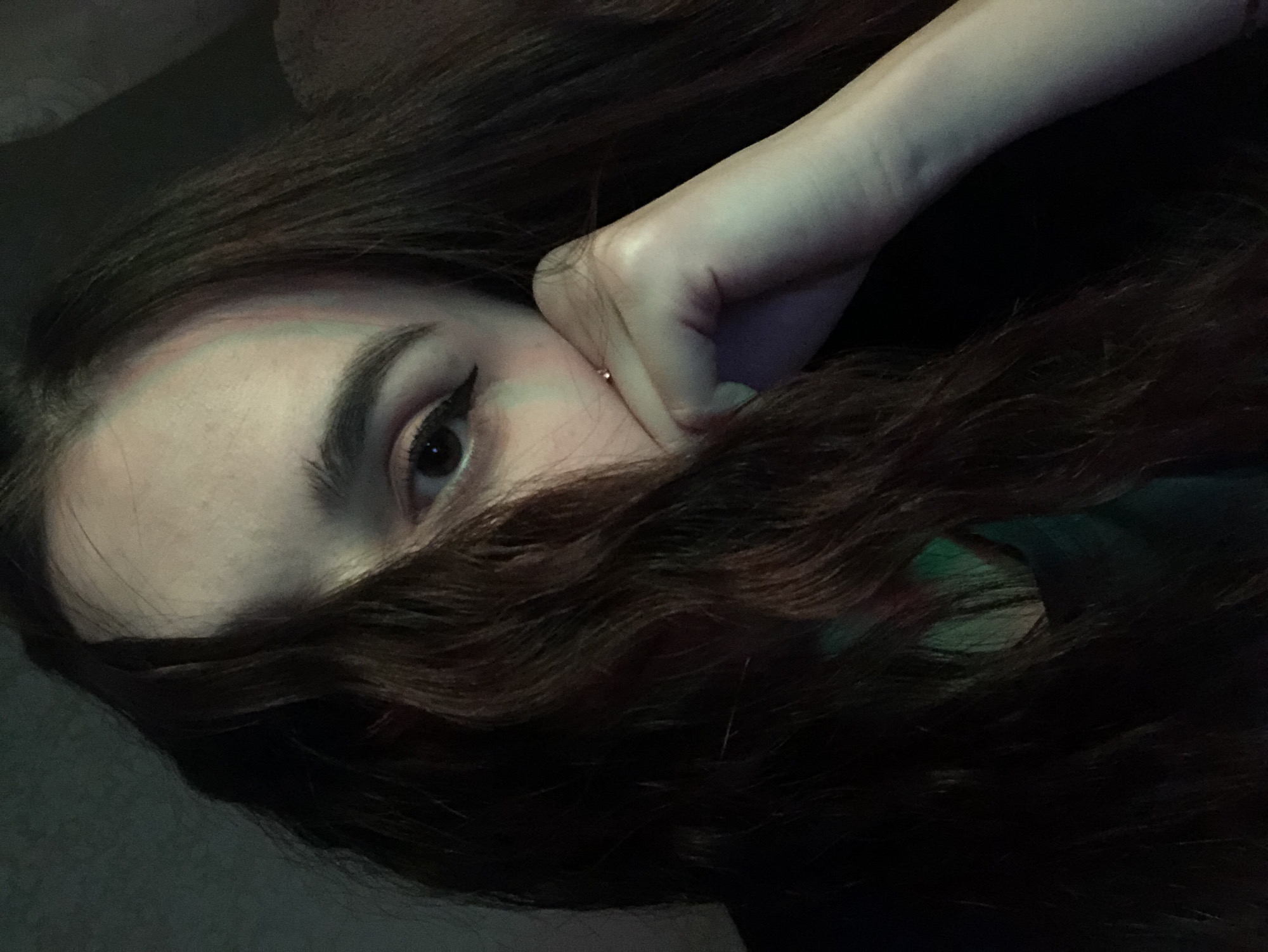 A close-up image of a person lying down, with their head turned to the side. The person has fair skin, dark hair, and one eye visible, featuring dark eyeliner. Their hand is positioned near their face, and hair partially covers the lower half of their face. The background is dark and indistinct, but hints of fabric suggest the person is lying on a couch or bed. The overall lighting is dim, casting shadows across the scene. There is no information to suggest the location is in the city of Noviy, Russia.
