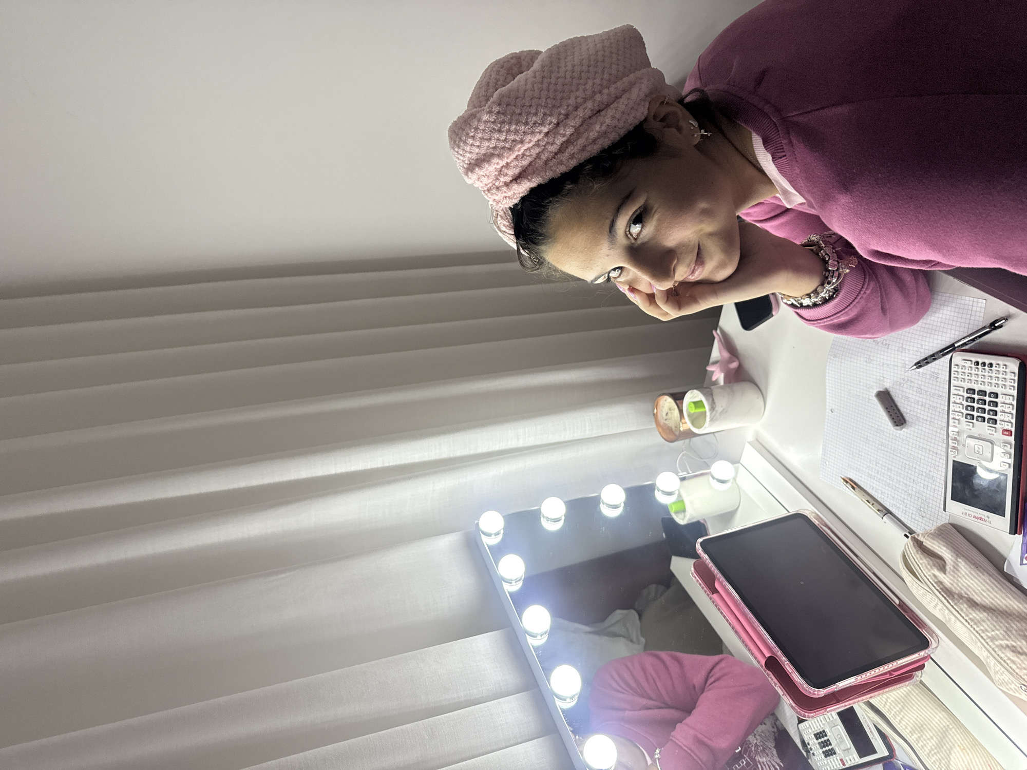 A young woman with dark hair wrapped in a pink towel sits at a vanity table with a lighted mirror. She is wearing a pink sweatshirt. The table contains a tablet, calculator, pen, and other miscellaneous items. The background shows white window blinds. There is no evidence to confirm the location as Vila Nova de Gaia, Portugal.