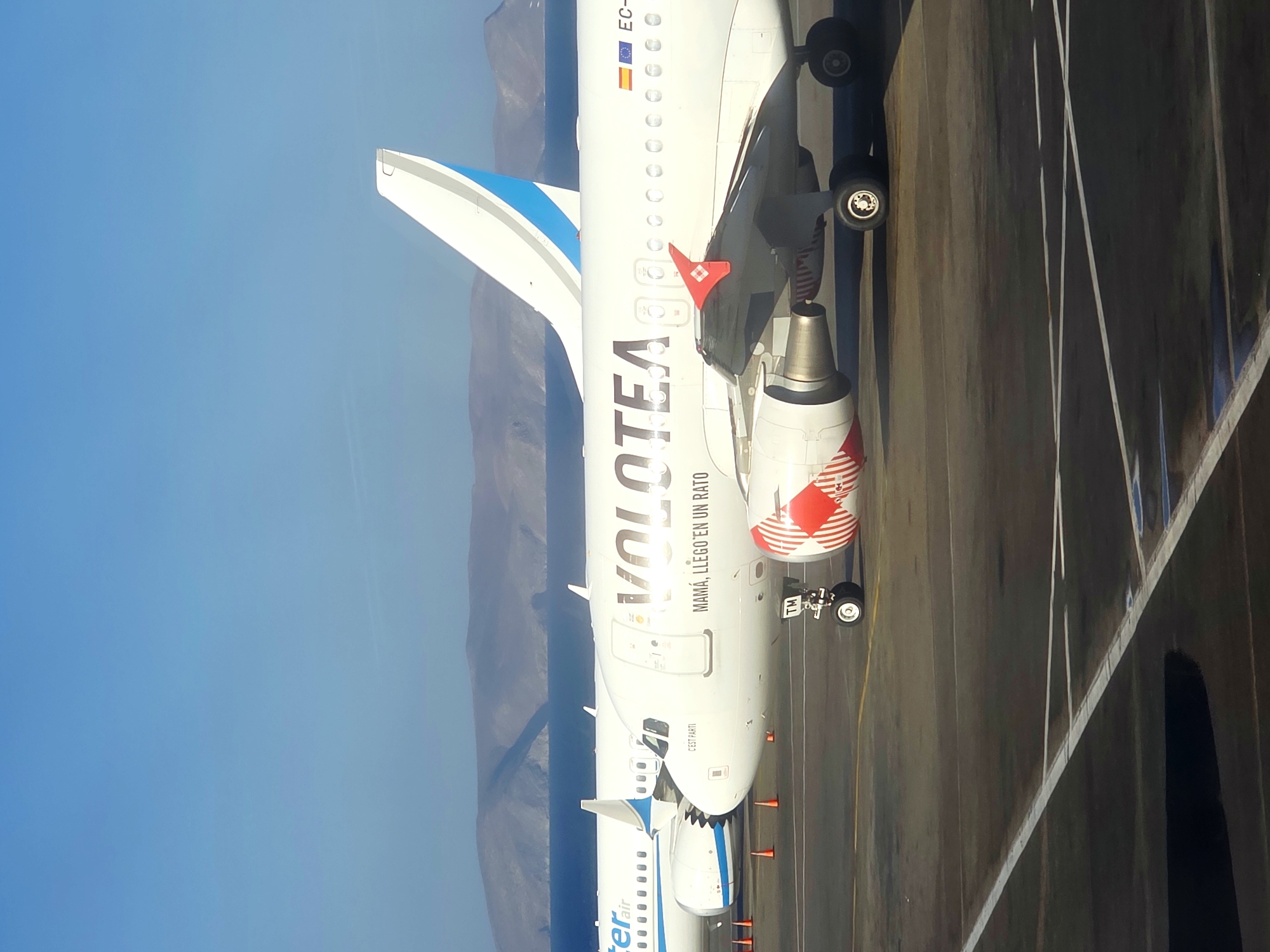 An exterior, eye-level, medium shot captures two white passenger airplanes parked on an asphalt tarmac under a clear blue sky. In the foreground, a Volotea airplane is positioned on the right, its engines featuring red and white stripes. The Volotea logo is visible on the fuselage, along with the text "MAMÁ, LLEGO EN UN RATO" and "C'EST PARTI". The Spanish flag and EU flag are also visible on the plane. To the left of the Volotea plane, partially obscured, is another white airplane with blue accents on its tail, with the word "ter air" visible on its fuselage. In the background, a body of water separates the tarmac from a range of hills or mountains. Several orange traffic cones are present on the tarmac between the two aircraft. The lighting suggests a sunny day. The location is identified as Nea Alikarnassos Municipal Unit, Greece.