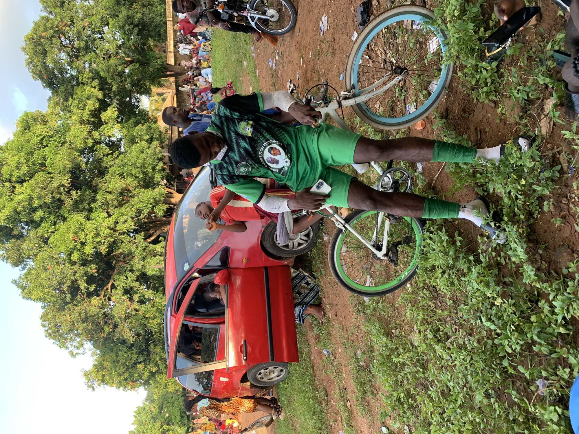 The image depicts a scene in Akwanga, Nigeria. A red car is parked with its driver's side door open. A man in a green soccer jersey and shorts is standing next to a bicycle, holding a phone. The bicycle has white frames and green and blue tires. The ground is dirt with patches of green vegetation. In the background, a crowd of people is visible under a tree. Another bicycle and a motorcycle are partially visible in the background as well.