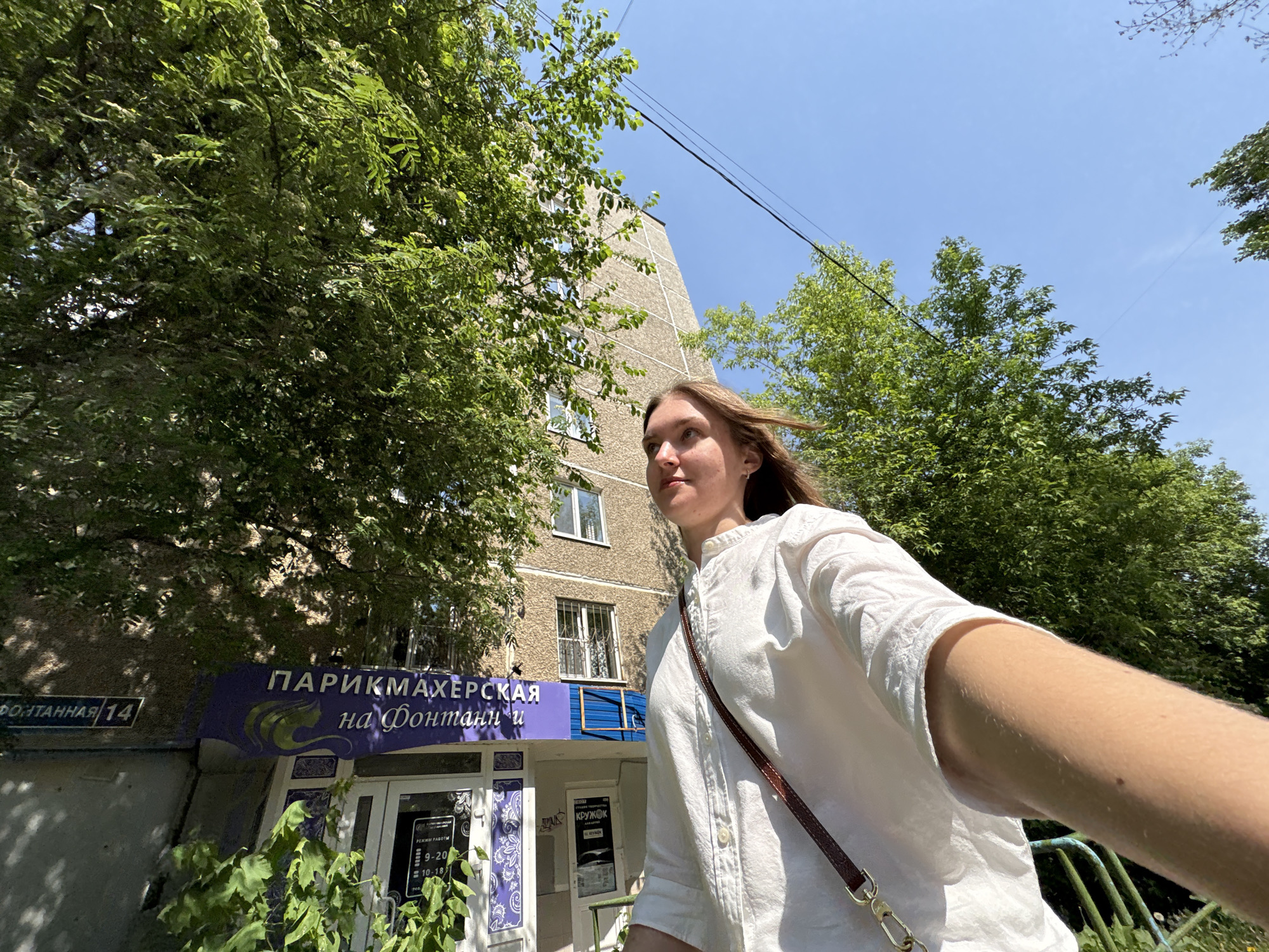 A woman is holding the camera and taking a selfie outdoors in Perm, Russia. The building behind her is a beauty salon with a sign in Russian, "ПАРИКМАХЕРСКАЯ на Фонтанн." The address "Фонтанная 14" is also visible on the building. She is wearing a white shirt and a brown purse with a golden chain strap. Trees surround the building. The sky is blue with some clouds.