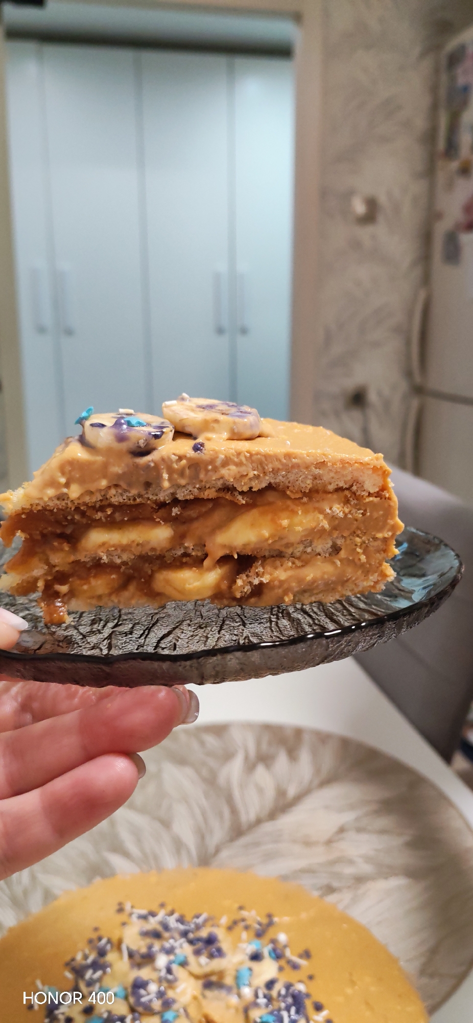 A person is holding a slice of caramel-covered layered cake on a textured glass plate. The cake appears to have banana slices between the layers and on top, with small purple and blue sprinkles as garnish. The background includes a white cabinet with four doors, light-colored wallpaper, and what appears to be a refrigerator. The location is not specified. A "HONOR 400" watermark is visible in the bottom left corner.