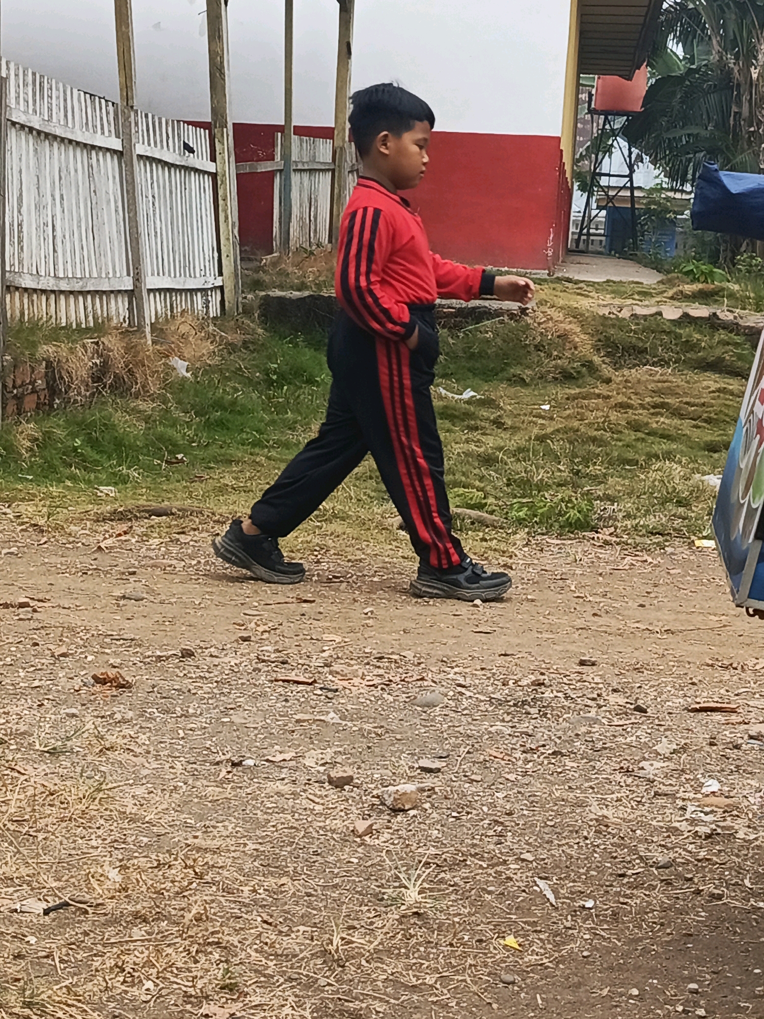 A young boy in a red and black tracksuit walks on a dirt path in front of a weathered white corrugated metal fence and a red building. He is wearing black athletic shoes. The path is covered in loose dirt and small pebbles, with patches of dry grass and weeds growing alongside. In the background, a large tree is visible, along with a blue tarp or covering. The sky is overcast and bright. This scene appears to be in an outdoor area in the city of Megang Sakti II, Indonesia.