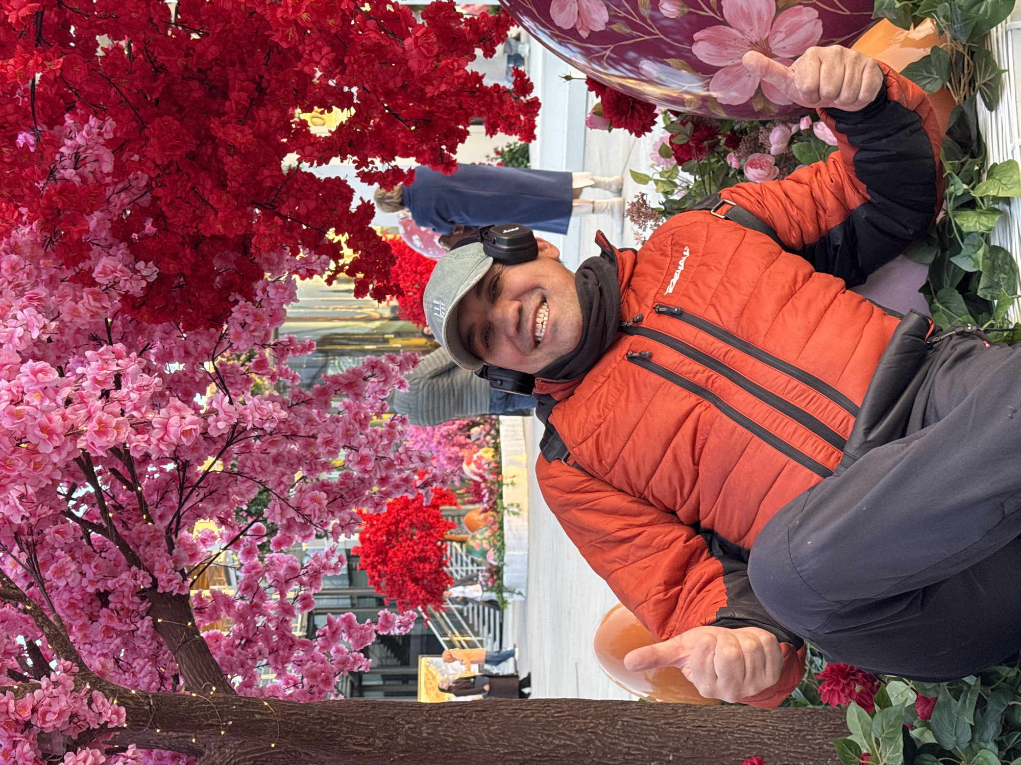 The image shows a smiling man in an orange jacket and a grey baseball cap with headphones around his neck, giving a thumbs-up. He is positioned in front of a large display featuring cherry blossoms in pink and red. The background suggests an indoor setting, possibly a shopping center in Moscow, Russia, given the presence of decorative Easter eggs and other floral arrangements. Other people can be seen in the background.