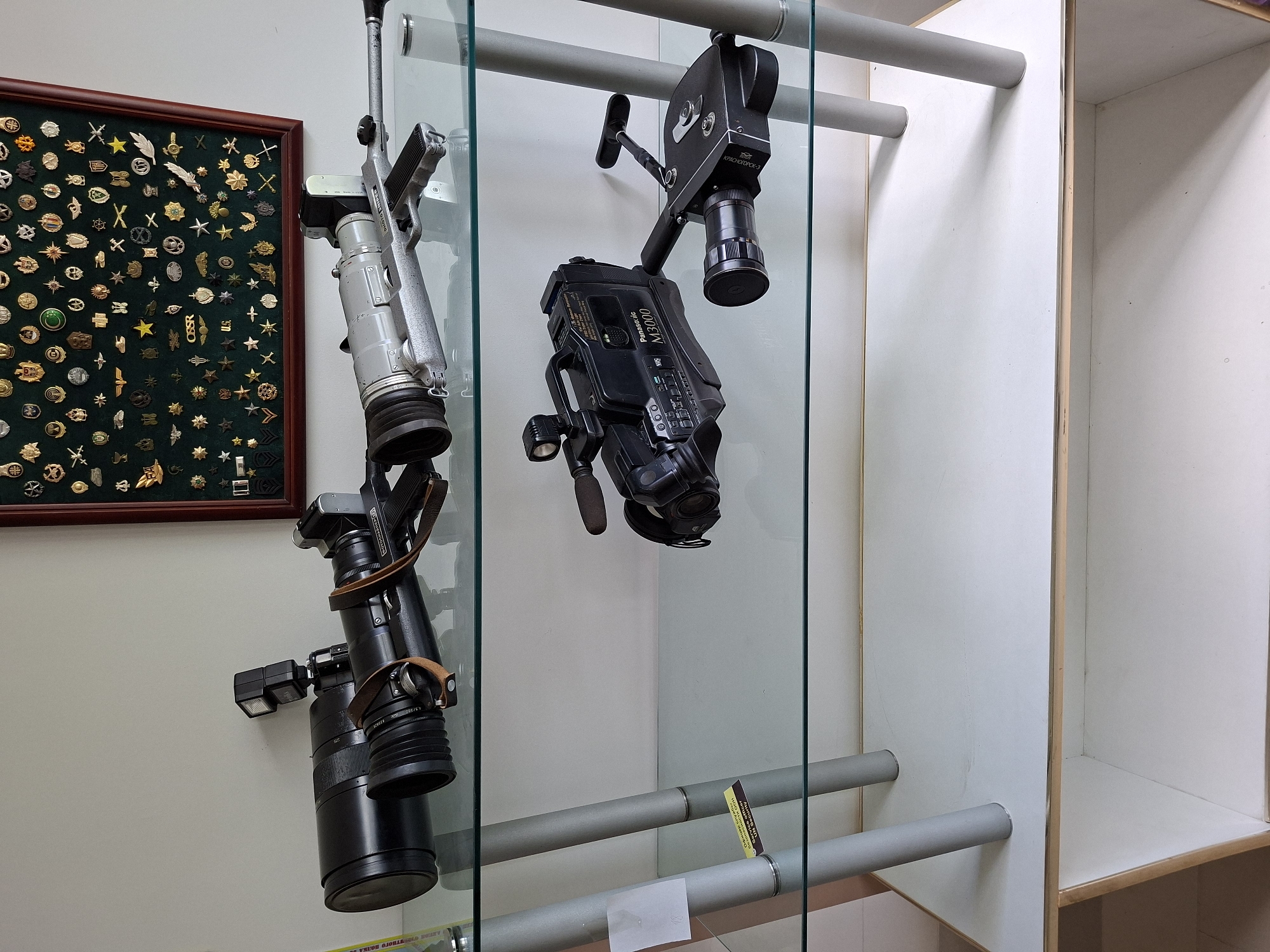 The image shows a glass shelf displaying various vintage cameras and photographic equipment. On the top shelf, there are two large cameras with long lenses. One is black with a flash unit attached, and the other is silver with a gun-like grip. Behind them, a framed display case filled with numerous medals and pins is mounted on the wall. The bottom shelf holds a black Panasonic M3000 video camera and a smaller, dark-colored camera with a lens and a handle. The scene appears to be indoors, in what seems to be a display or storage area. The location is identified as Ialoveni, Moldova.
