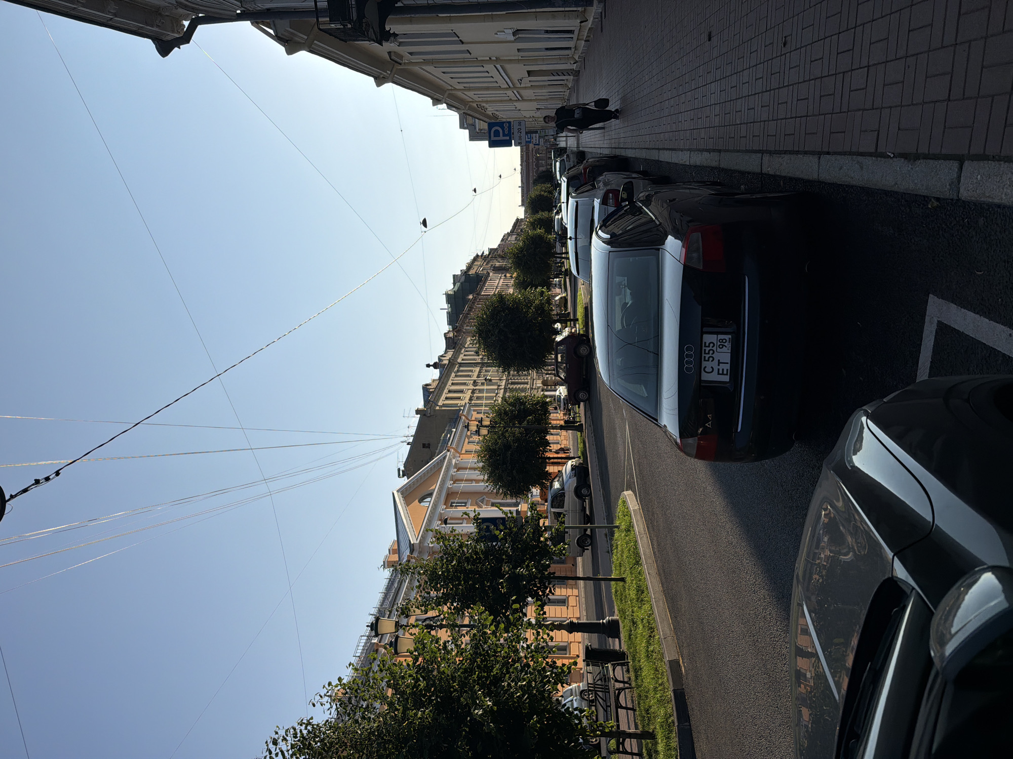 The image captures a street scene in Saint Petersburg, Russia, likely in the late afternoon or early evening based on the long shadows and the warm glow of the setting sun on the buildings. The perspective is from street level, looking down a wide avenue lined with classical European architecture, predominantly painted in shades of orange and yellow. The buildings feature ornate facades with columns and detailed cornices.
On the left side of the street, a sidewalk area is visible with mature trees and lampposts. A few cars are parked along the curb. The road itself is asphalt, and further down, more cars are parked on both sides of the street.
On the right side of the street, a pedestrian sidewalk paved with red bricks is prominent. A person is walking away from the camera, carrying a dark bag. A blue and white parking sign with Russian text is affixed to a pole on this side.
Several cars are parked along the right curb. The most prominent car in the foreground on the right is a dark sedan, possibly an Audi, with a license plate visible. In the foreground on the left, the rear end of another dark car is visible.
Overhead, numerous thin wires crisscross the sky, characteristic of urban infrastructure. The sky is a clear, pale blue, suggesting a pleasant day. The overall impression is one of a tranquil urban environment during a quieter time of day.