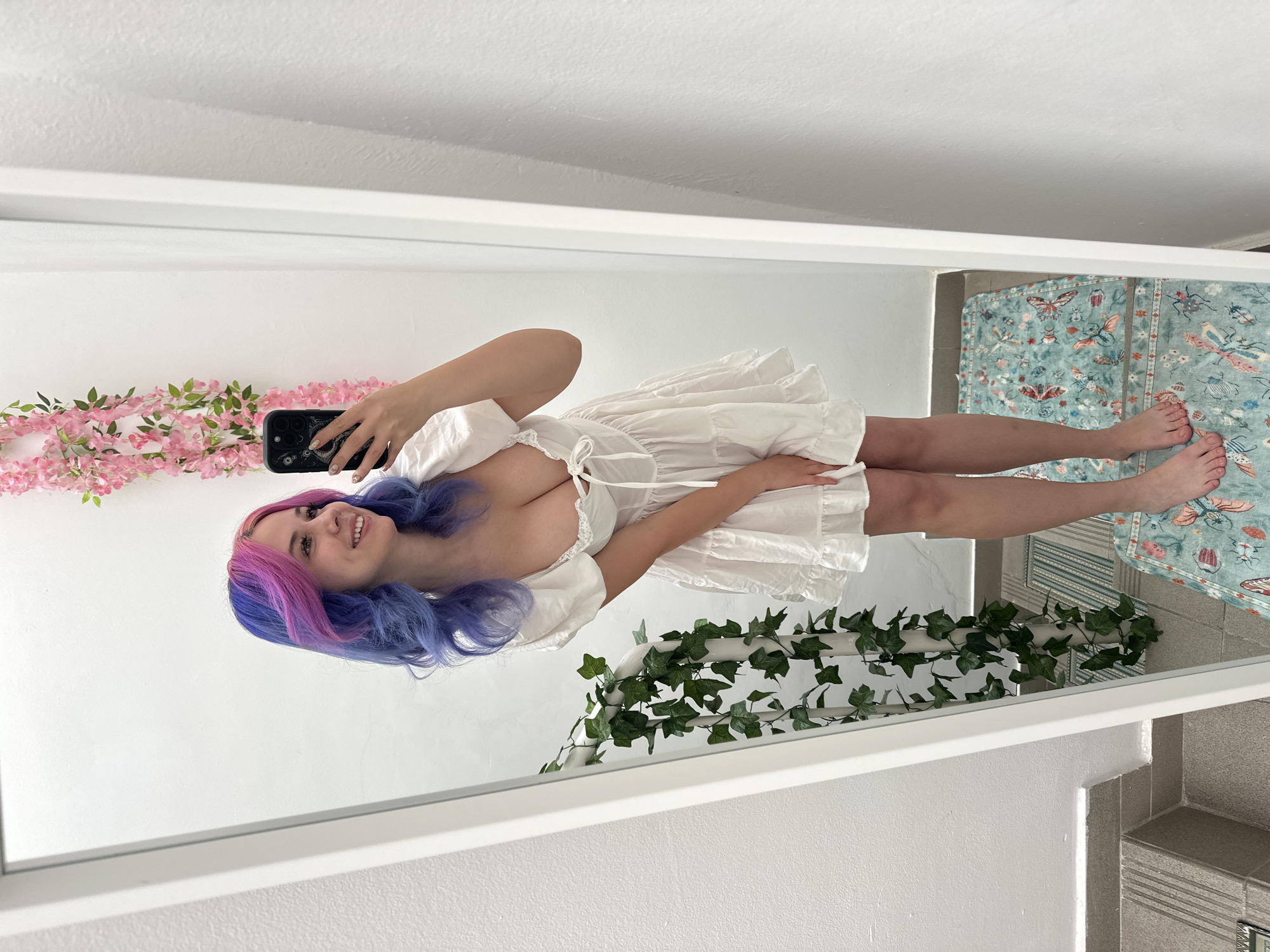 The image is a mirror selfie featuring a young woman with long, wavy hair dyed in vibrant purple and pink hues. She is wearing a short, white, off-the-shoulder dress with a gathered bodice and a ruffled skirt. She holds a dark-colored smartphone, possibly an iPhone, in her right hand to take the photo. Her bare feet are visible on the patterned rug at the bottom right.
The woman is smiling cheerfully, looking directly into her phone's camera reflection. The background visible in the mirror reflection includes a plain white wall. Decorative elements frame the scene: a string of artificial pink cherry blossom-like flowers is positioned on the left side, and a green artificial ivy garland is draped over a white rail or shelf on the right. The floor in the reflection shows light grey tiles, partially covered by a light blue rug or mat adorned with a detailed pattern of butterflies and other insects in various colors. A grey tiled structure is also discernible on the far right. The scene is brightly lit, indicative of an indoor setting. There is no visible text in the image, nor are there any discernible visual cues to specifically identify the location as Zagreb, Croatia, from the image content itself.