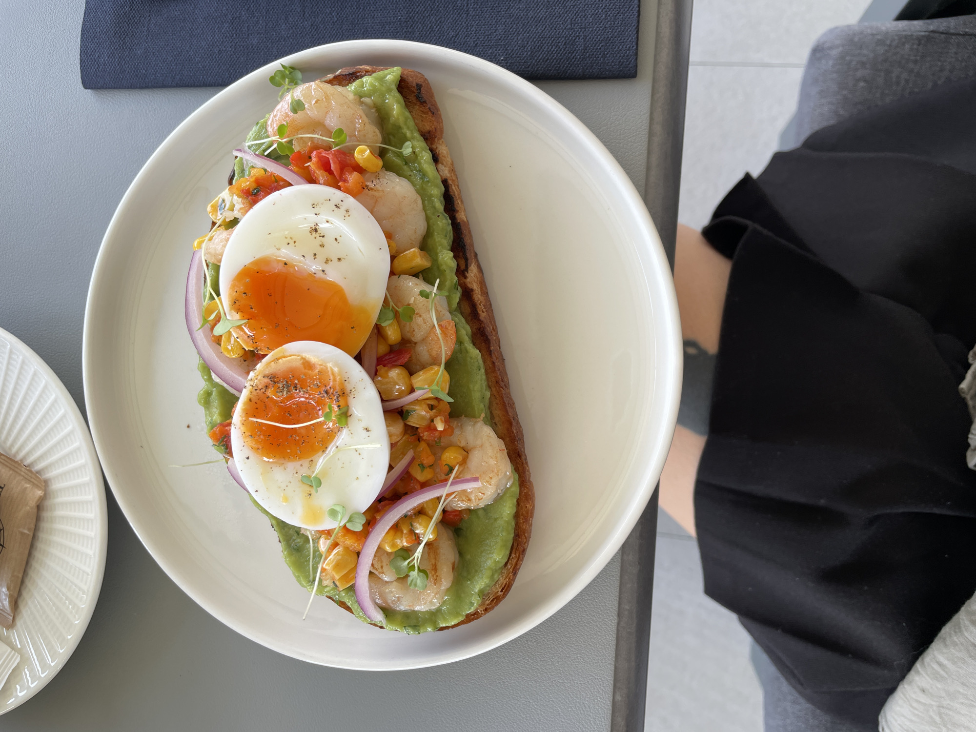 A top-down view reveals a plate of food, an adjacent plate, and a portion of a person seated at a table.
The main focus is a white plate containing a slice of toast generously spread with mashed avocado. Toppings include cooked shrimp, corn, red bell pepper, thin slices of red onion, microgreens, and two soft-boiled eggs seasoned with pepper.
To the left of this plate is a smaller white plate with an embossed pattern, holding a small paper packet.
To the right of the plates, part of a person is visible. They are wearing dark clothing, possibly black pants or a skirt, with a lighter-colored garment partially visible at the edge. A glimpse of a tattoo is visible on their leg.
The setting appears to be a restaurant or cafe, possibly outdoors, suggested by the grey surfaces and the overall lighting. The location in the city of Boreti, Montenegro is not discernable from the visual information.