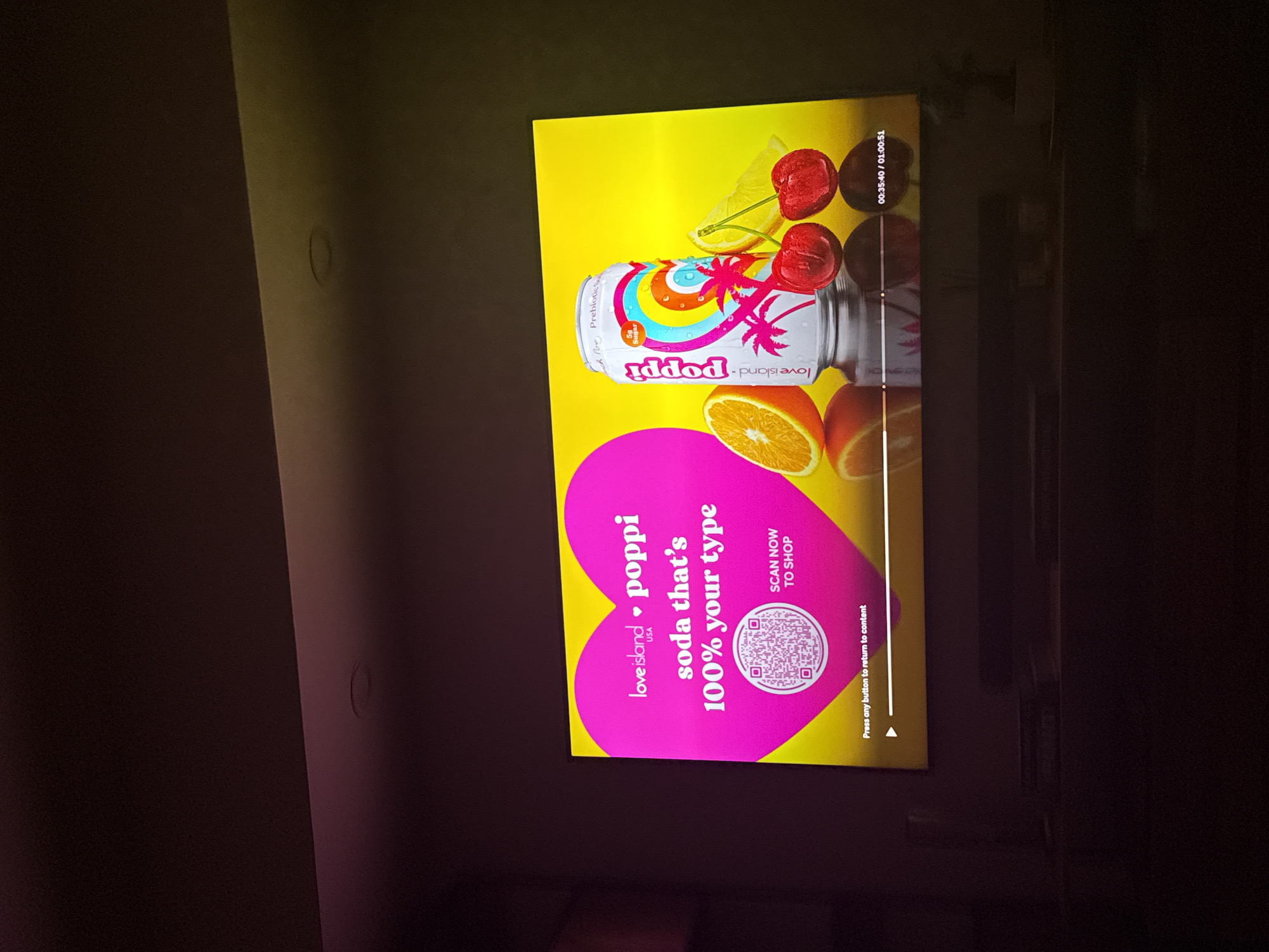 The image displays a television screen showing an advertisement for "Love Island USA" and "Poppi" soda. The advertisement features a large pink heart with the text "loveisland USA · poppi soda that's 100% your type" inside. A QR code is visible within the heart, with the text "SCAN NOW TO SHOP" below it. To the right of the heart, there is a can of Poppi soda with a colorful design, alongside an orange slice, a lemon wedge, and two cherries. The background of the advertisement is bright yellow. The bottom of the screen shows a progress bar with timestamps "00:35:40 / 01:00:51" and the text "Press any button to return to content". The overall scene is dark, indicating the room is dimly lit. There are no people visible in the image, and no specific location in the city of Miami, United States of America, can be determined from the visual information provided.