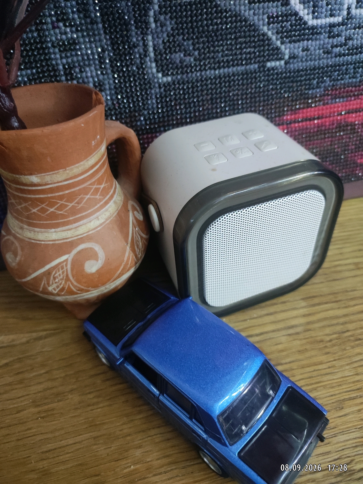 An overhead medium shot shows a rustic ceramic jug with white decorations, a white portable speaker with a grey trim, and a blue toy car. The objects are arranged on a wooden surface, with a dark, textured background that appears to be fabric. The jug is on the left, tilted slightly, and has a handle. The speaker is in the center, facing the viewer. The toy car is in the foreground, in front of the jug and speaker, with its front facing right. The image is dated 08.09.2026 at 17:28.