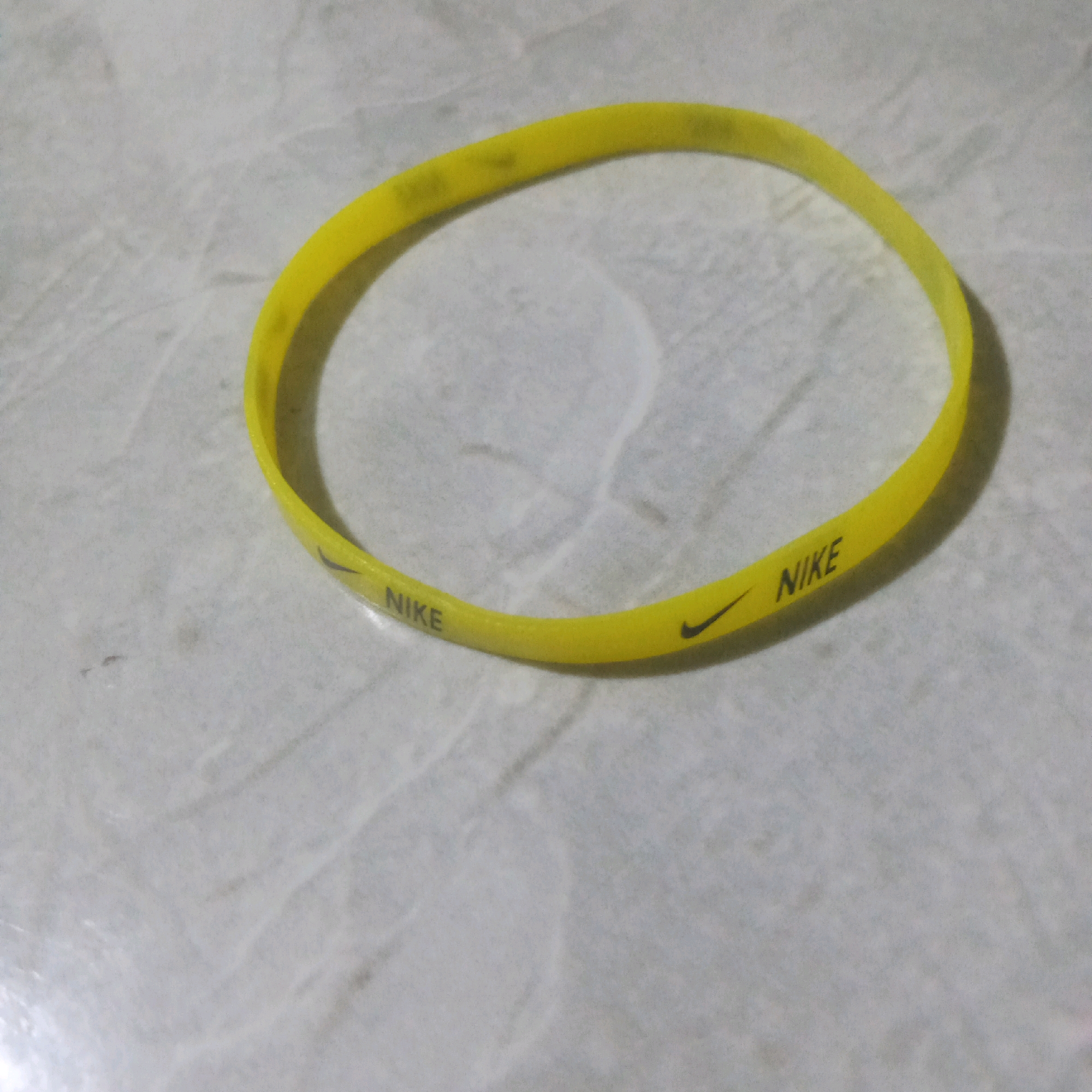 A yellow Nike branded wristband is lying on a mottled gray surface. The wristband has "NIKE" printed on it in black, along with the Nike swoosh logo. The surface appears to be a countertop or table with subtle variations in color and texture. The image is taken from a slightly elevated angle, with the wristband forming a loose curve across the frame. The background is out of focus, but the marbled pattern of the surface is visible. There is no indication of any specific location within Kendayakan, Indonesia, as the image solely depicts the wristband and its immediate surroundings.