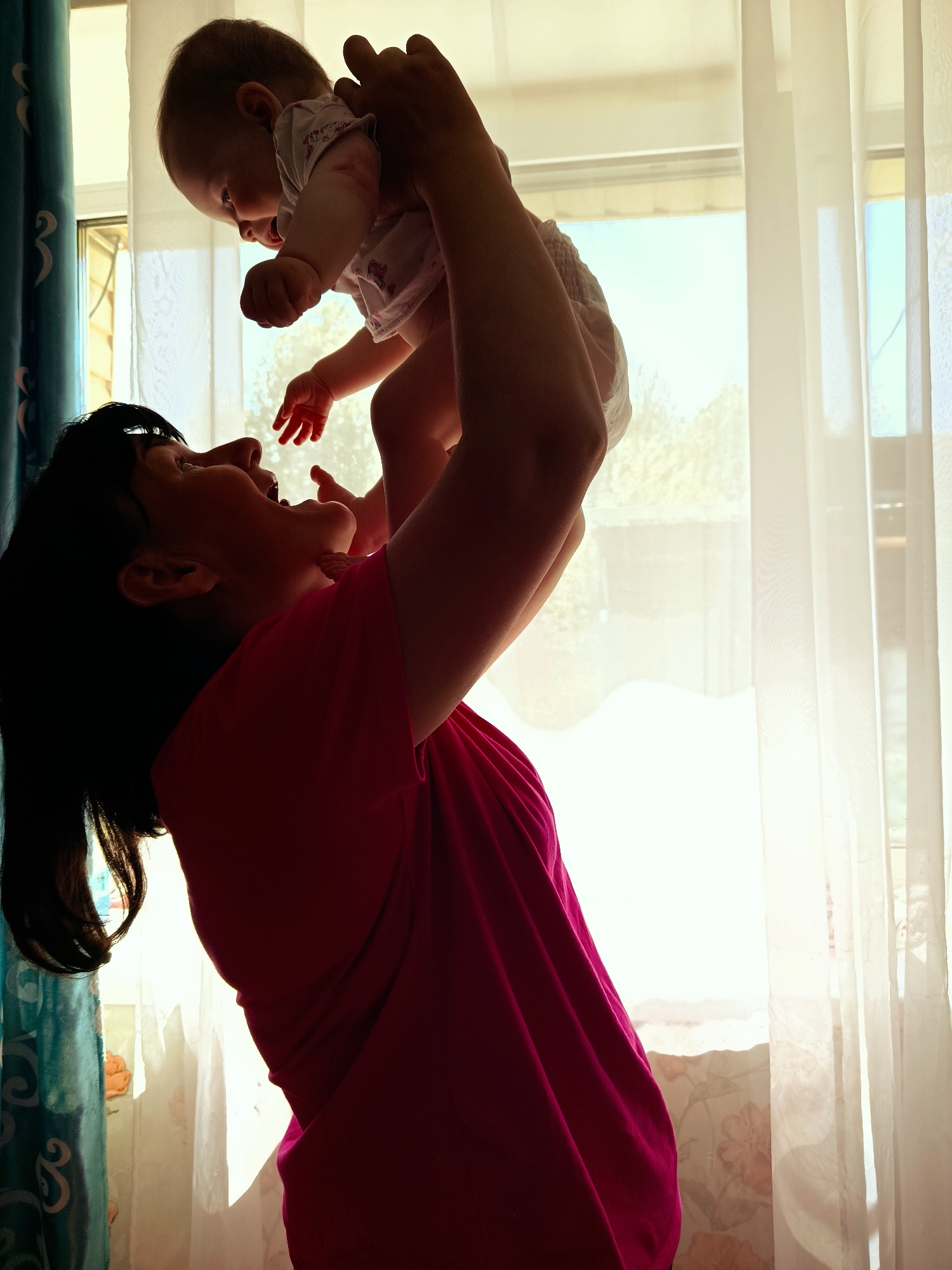 A woman in a pink shirt is holding a baby up in the air. They are near a window with white curtains. There is a floral patterned wallpaper and a blue patterned curtain in the background. The location appears to be in a home. It is day and there is a bright light coming through the window.