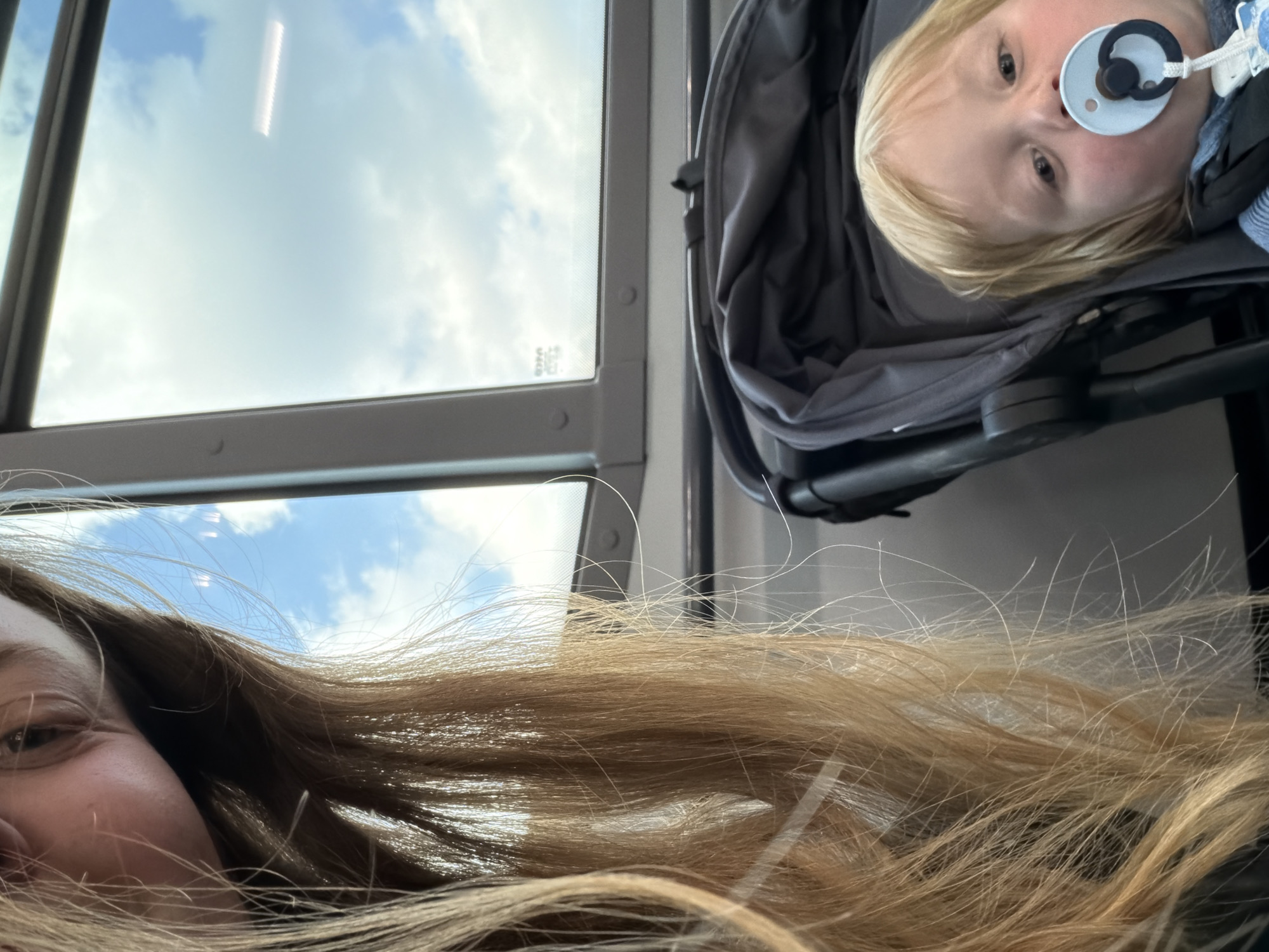 The image is captured from a low, upward-looking perspective, presenting a rotated view of the scene. It features two individuals and an expansive view of the sky through a window, suggesting travel within a vehicle.
In the foreground, the bottom half of the image is dominated by long, light brown hair belonging to an adult. Part of this adult's face, specifically an eye and cheek, is visible on the bottom left, showing a hint of a relaxed expression or smile.
To the right and above the adult's hair, a baby or toddler is seen lying in a dark grey stroller or car seat. The child has blonde hair, light eyes, and a white and black pacifier in their mouth, looking upwards with a calm expression. They are dressed in a blue and white striped top beneath a darker layer.
The upper and left portions of the frame are occupied by a window, framed in dark grey, which reveals a bright blue sky dotted with scattered white clouds. The window structure suggests the interior of a train, bus, or similar public transport vehicle. The clear sky indicates it is daytime.
No discernible text is visible within the image. The scene depicts a quiet moment of travel, likely a parent and child, in transit under clear daytime skies, with the location context being Solzago, Italy.