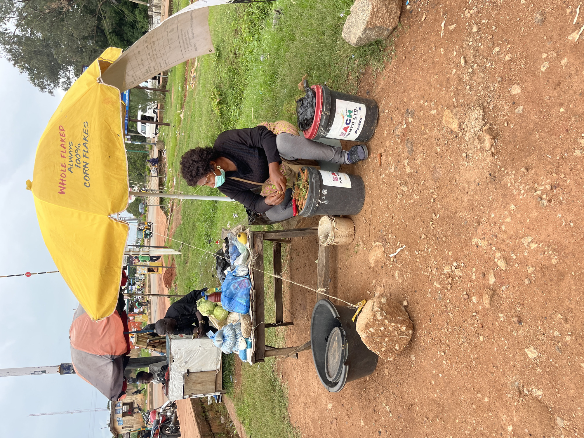 In Bukuru, Nigeria, a woman wearing a face mask sits beside a market stall under a yellow umbrella. The umbrella has "WHOLE FLAKED ALWAYS 100% CORN FLAKES" written on it. The woman is wearing a dark long-sleeved shirt, grey pants, and sandals. She is sorting through some vegetables or produce, with a bucket of what appears to be carrots next to her.
The market stall itself is a wooden table laden with various goods. These include bags of what looks like rice or beans, cabbages, and bottles of liquid. Two large black buckets are visible in the foreground, one containing a black bag and the other with a label for "EACH PAINTS LTD. Putty". A large grey bucket with a lid sits to the left, and a large rock is placed in front of it.
In the background, other market activity can be observed. A man in a black outfit is seated near another stall, and motorcycles are parked. A smaller stall with an orange and grey umbrella is also visible. The ground is a reddish-brown dirt, with patches of green grass and various rocks scattered around. The sky is overcast. The overall scene depicts a typical day at an outdoor market in Nigeria.