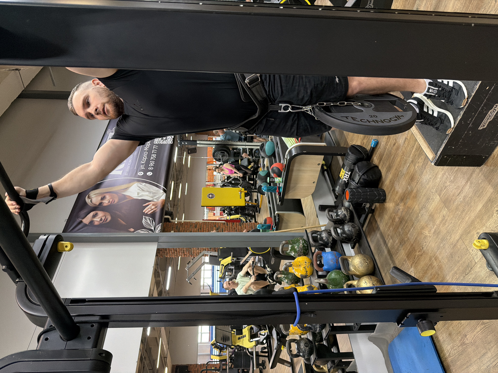 Here is a detailed description of the image:
**Scene:**
The image depicts the interior of a gym in the city of Chaikovsky, Russia. The camera angle is slightly tilted.
**People:**
- A man in black athletic wear is performing a hanging exercise. He has a weight plate attached to a belt around his waist. He has a beard and is holding onto handles of a machine.
- Other people are indistinctly visible in the background, presumably also exercising.
**Objects:**
- Weight lifting equipment (various machines, kettlebells, weights)
- A "Technogym" weight plate
- TRX cables
- A sign indicating the location as "ул. Карла-Марк" and a phone number.
**Actions:**
The man is performing a hanging exercise, likely a variation of a pull-up or leg raise.
**Interactions:**
The man is interacting with the gym equipment. Other people are likely interacting with the gym equipment, but their actions are not clear.
**Environment:**
The gym has a wooden floor and appears to be well-lit.