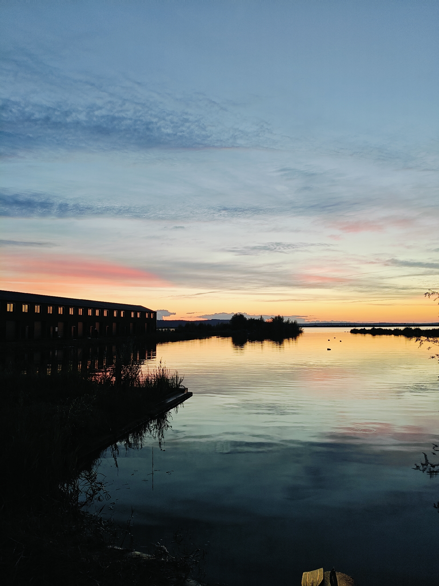 The image depicts a serene sunset scene over a body of water, likely a reservoir or large river, in Chaikovsky, Russia. In the foreground to the left, dark silhouettes of reeds and grasses line the edge of the water, partially obscuring a wooden platform extending into the water. The water itself is calm, reflecting the colors of the sky and the structures along the shore.
Dominating the left side of the frame is a long, dark, multi-story building with numerous illuminated windows, suggesting it's either an apartment complex or a commercial property. The building's reflection is clearly visible on the still water. Further in the distance, across the water, are several small, dark, tree-covered islands.
The sky is a canvas of soft twilight colors. The upper portion is a pale blue, transitioning to streaks of delicate pink and orange near the horizon, indicative of sunset. Thin, wispy clouds are scattered across the sky, catching the fading light. The overall atmosphere is tranquil and peaceful, capturing the quiet beauty of the natural environment juxtaposed with human architecture. There are no people visibly present in the scene. The image is a still shot, implying no movement or action is occurring.