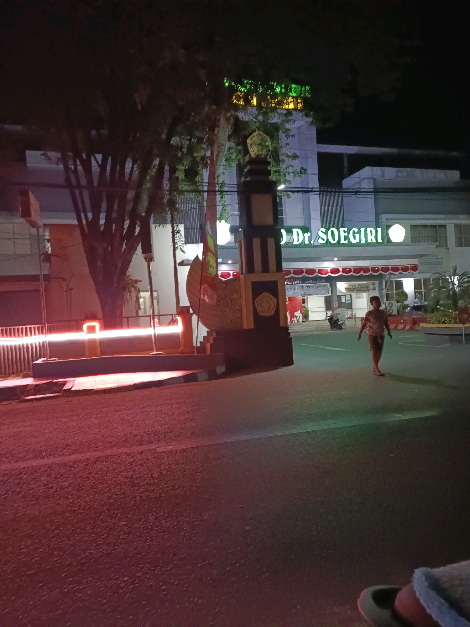 This image captures a nighttime street view of Dr. Soegiri Hospital in Lamongan, Indonesia. A young child is walking across the street in front of the hospital entrance. The hospital building features prominent green neon signage with the name "Dr. SOEGIRI". A decorative monument stands at the entrance. Red and white lights illuminate a barrier to the left of the frame, reflecting on the asphalt road. The scene is illuminated by artificial lights, creating a somewhat low-light environment.