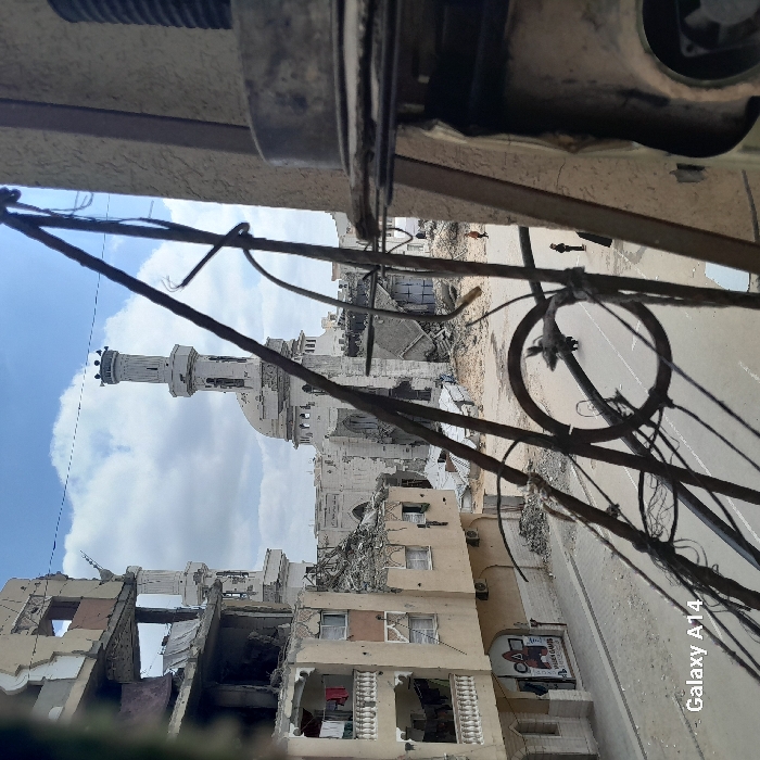 The image depicts a devastated urban street scene in Khan Yunis, Palestinian Territory, viewed from an elevated position, likely from within a damaged building. In the immediate foreground, distorted metal wires, rebar, and elements of a broken structure, including a circular metal component, frame the view on the right and center.
To the left, a multi-story residential building stands heavily damaged, with multiple floors exposed, concrete fractured, and rebar visible. Clothes hang on several balconies, suggesting recent occupancy or hurried abandonment. The building's facade shows numerous impact marks or bullet holes.
In the midground, a paved street, largely cleared but with scattered debris, runs horizontally across the frame. Three individuals are visible on the street: two closer to the left, one wearing an orange/brown top walking away, and a smaller figure in dark clothing. A third, less distinct figure is further down the street to the right.
Dominating the background is a large mosque, identifiable as the Great Mosque of Khan Yunis, which has sustained extensive damage. Its white minaret remains standing but shows signs of structural compromise, and the main dome and surrounding mosque structure are severely damaged, with large sections reduced to rubble or missing, exposing internal elements. The sky above is partly cloudy. The overall scene is characterized by widespread destruction and rubble. A "Galaxy A14" watermark is present in the lower-left corner.