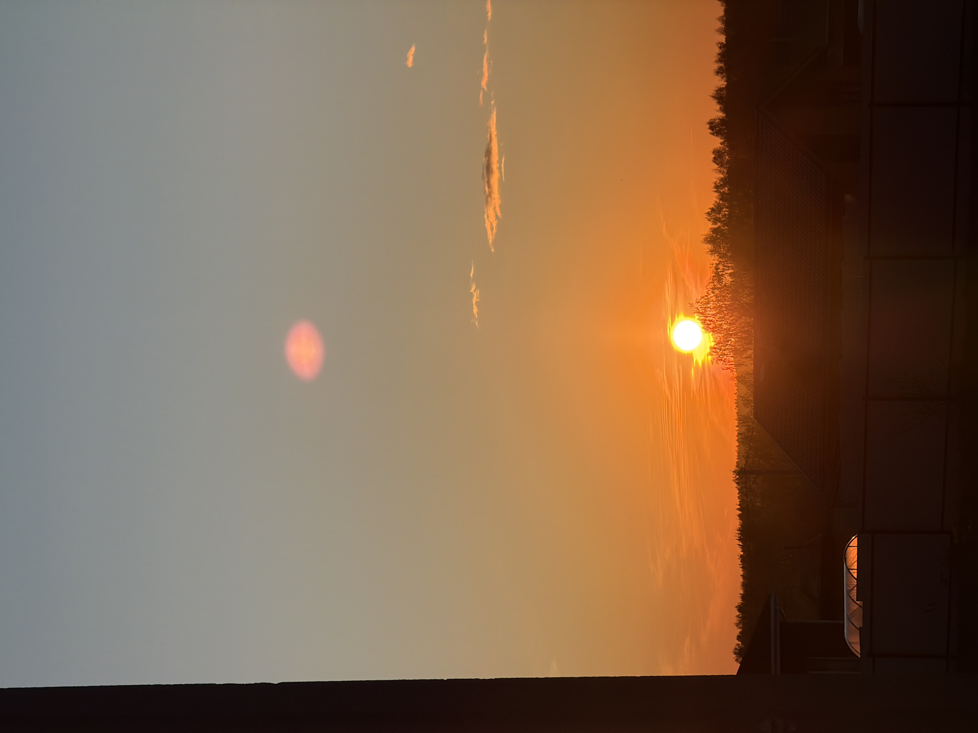 The image captures a sunset in Старые Кены, Russia. The sky transitions from blue on the left to orange towards the horizon where the sun is setting. Sparse clouds are visible in the sky. The horizon line is marked by a treeline and buildings. A structure with a curved roof is visible on the right. A dark surface occupies the bottom edge of the image.
